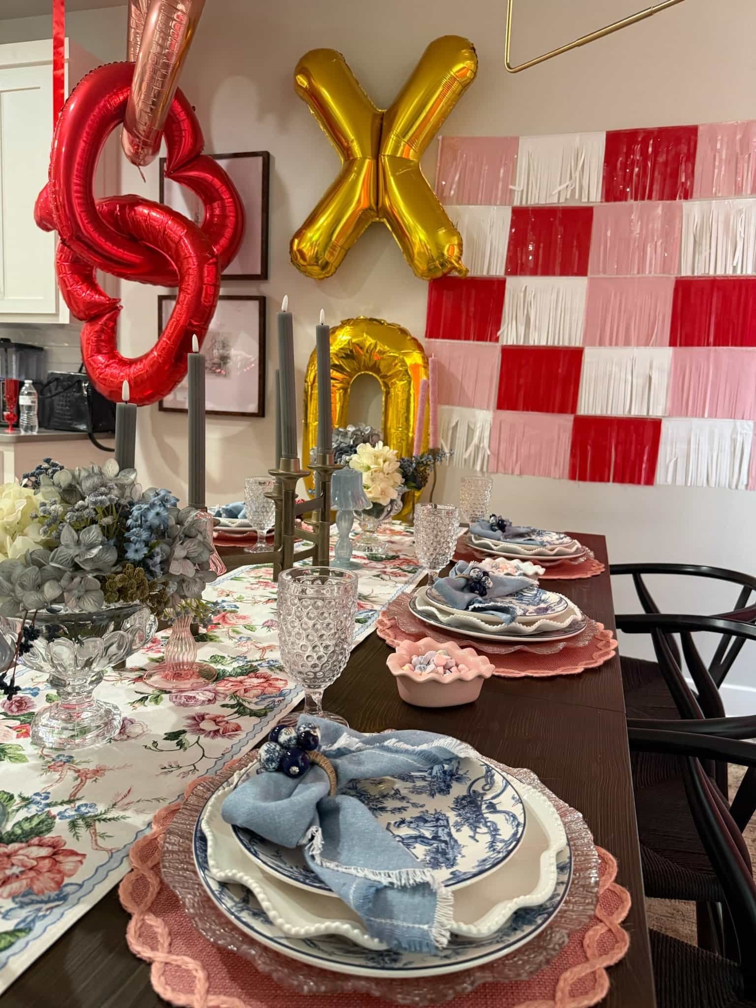 Festive table set with floral dishes, pastel napkins, candles, and XO balloons; red and pink wall decor in the background.