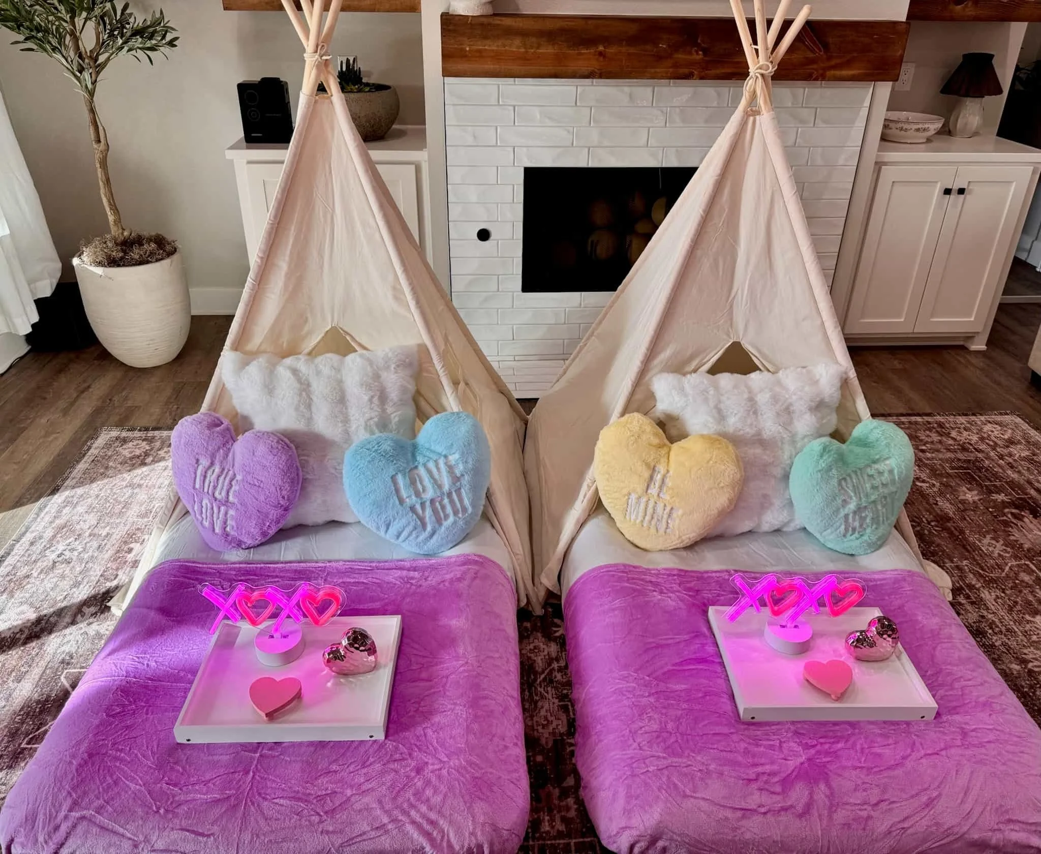 Two indoor teepees with purple bedding, heart pillows, and trays decorated with neon XOXO lights and pink hearts.