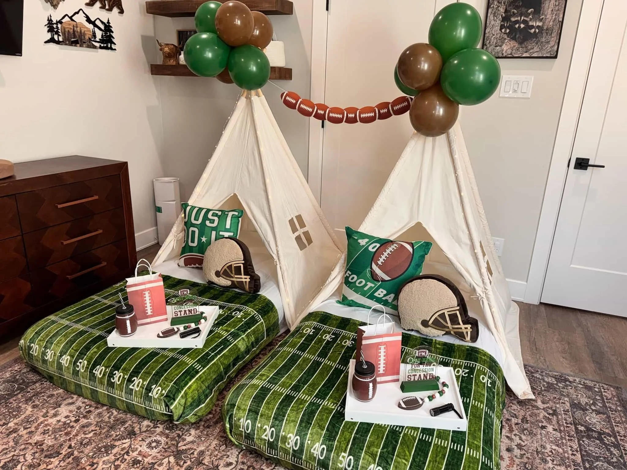 Two small tents with football-themed pillows and trays, decorated with balloons, set up for a football-themed indoor sleepover.
