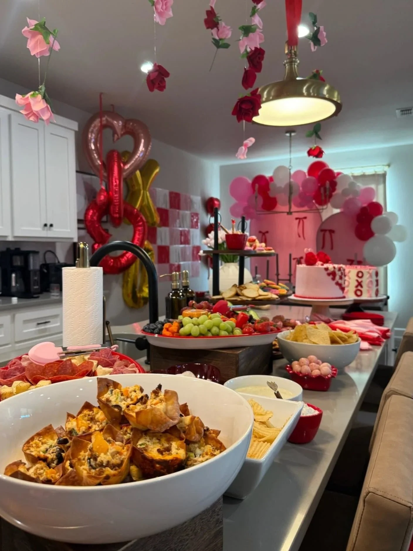 Valentine-themed party spread with balloons, flowers, desserts, fruit, and snacks on a kitchen counter.