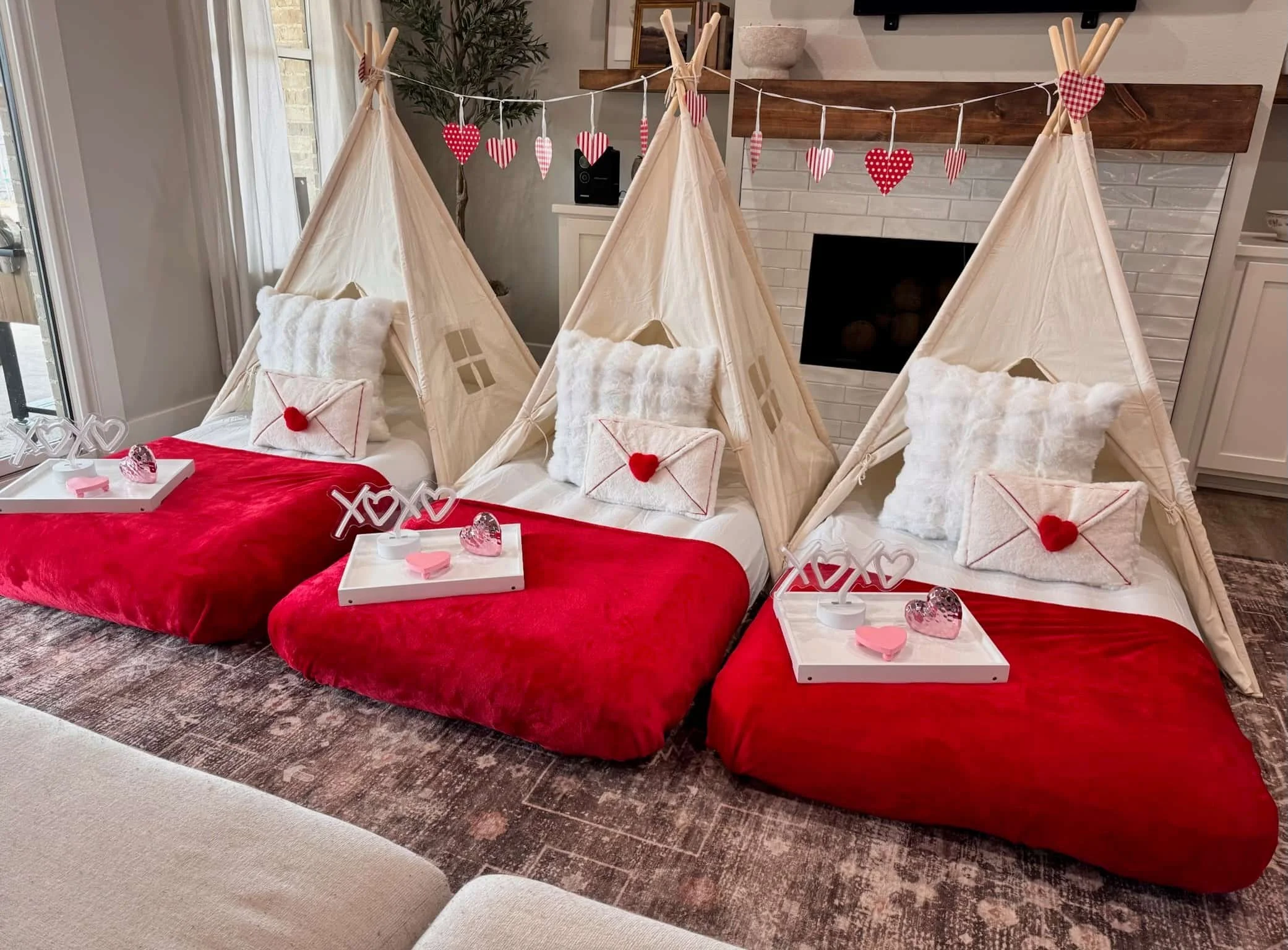 Three indoor teepees with red bedding, white pillows, and heart decorations, each with a tray holding Valentine treats.