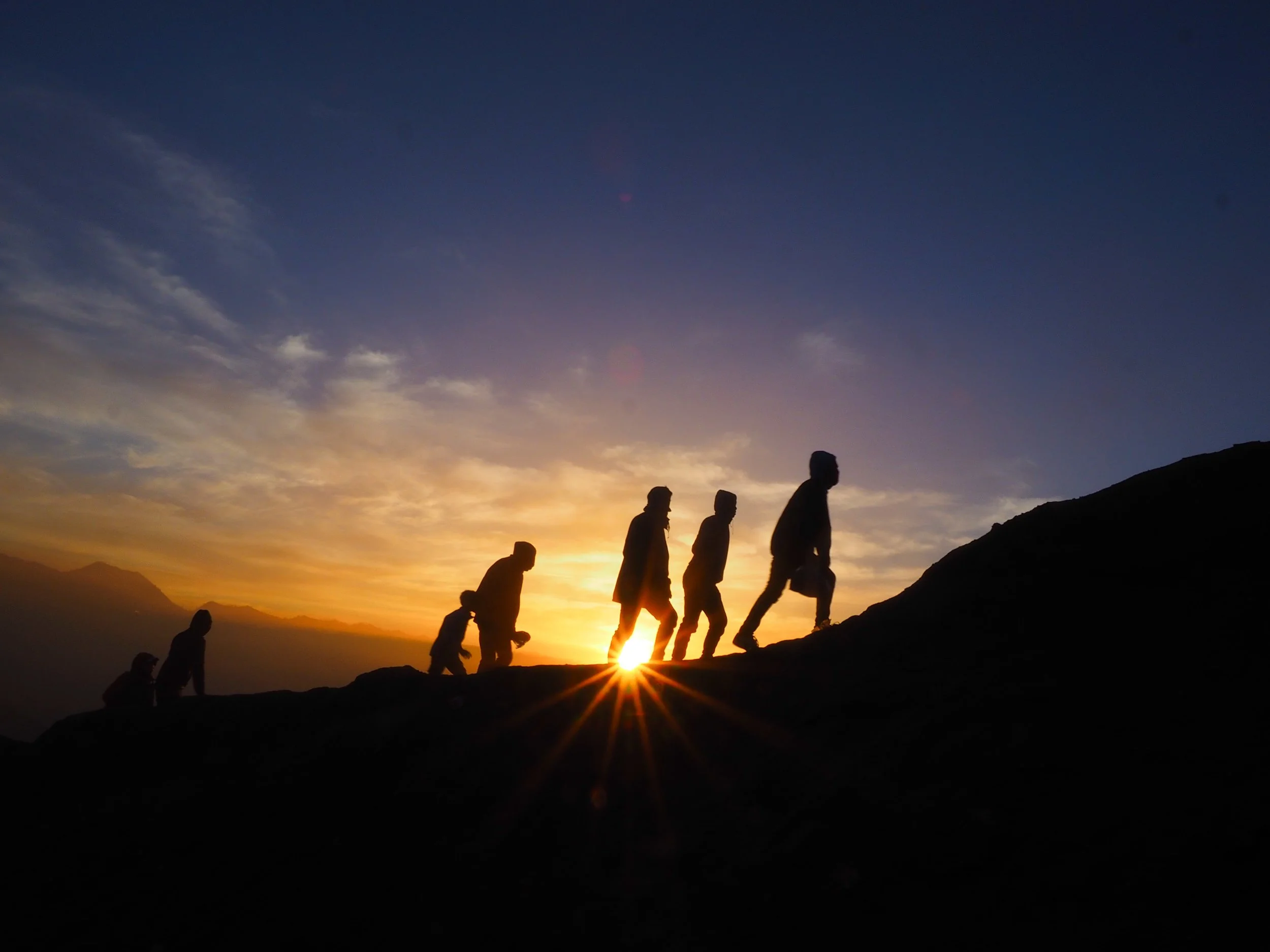Silhouetted group of hikers climbing a mountain during sunset with a colorful sky and mountain silhouettes in the background.