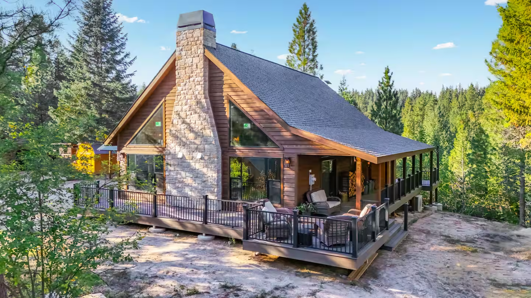A modern cabin house surrounded by tall evergreen trees, with a wooden deck and outdoor seating area, situated in a forested area on a clear day.