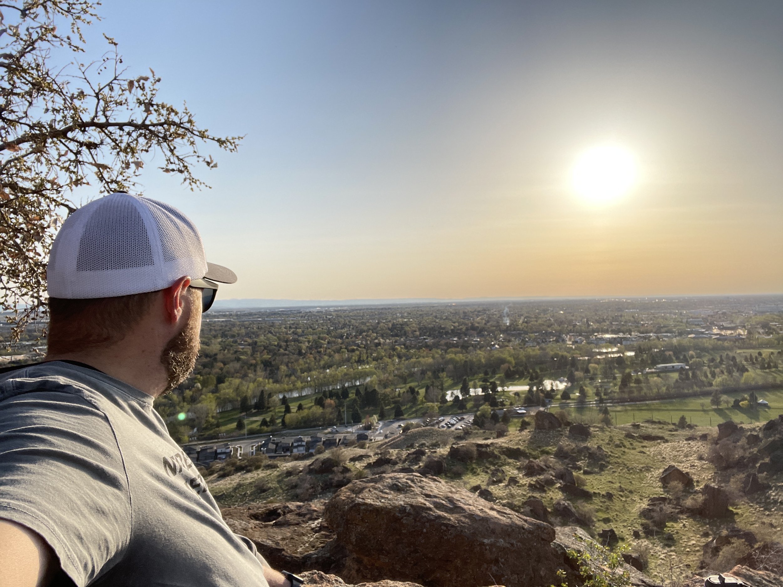 A man wearing a baseball cap and sunglasses looking at a sunset over a cityscape, with rocky terrain in the foreground and a tree partially visible on the left.