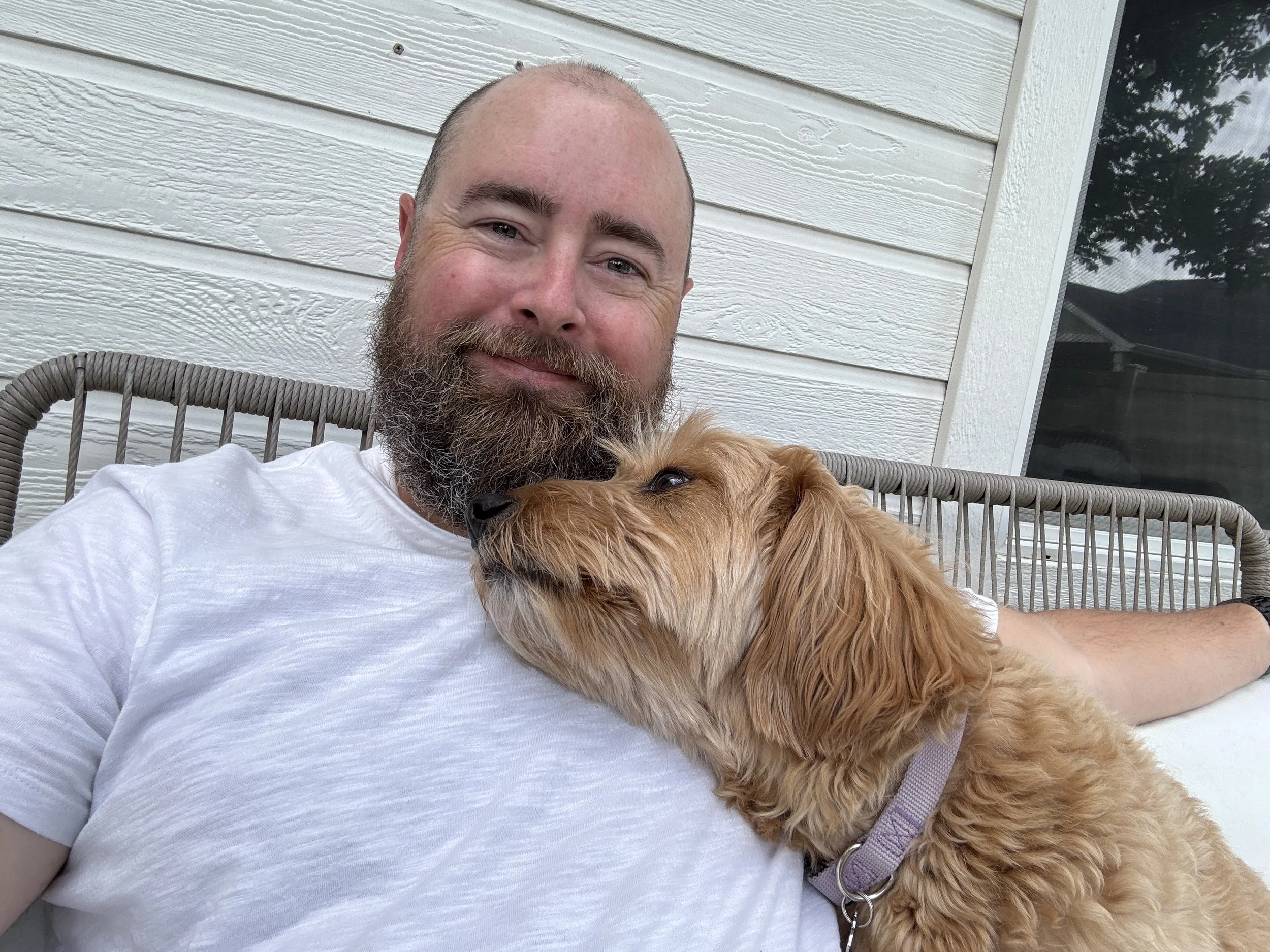 A man with a beard and a white t-shirt relaxing outside on a gray wicker chair, with a golden-colored dog resting its head on his chest, both looking content.