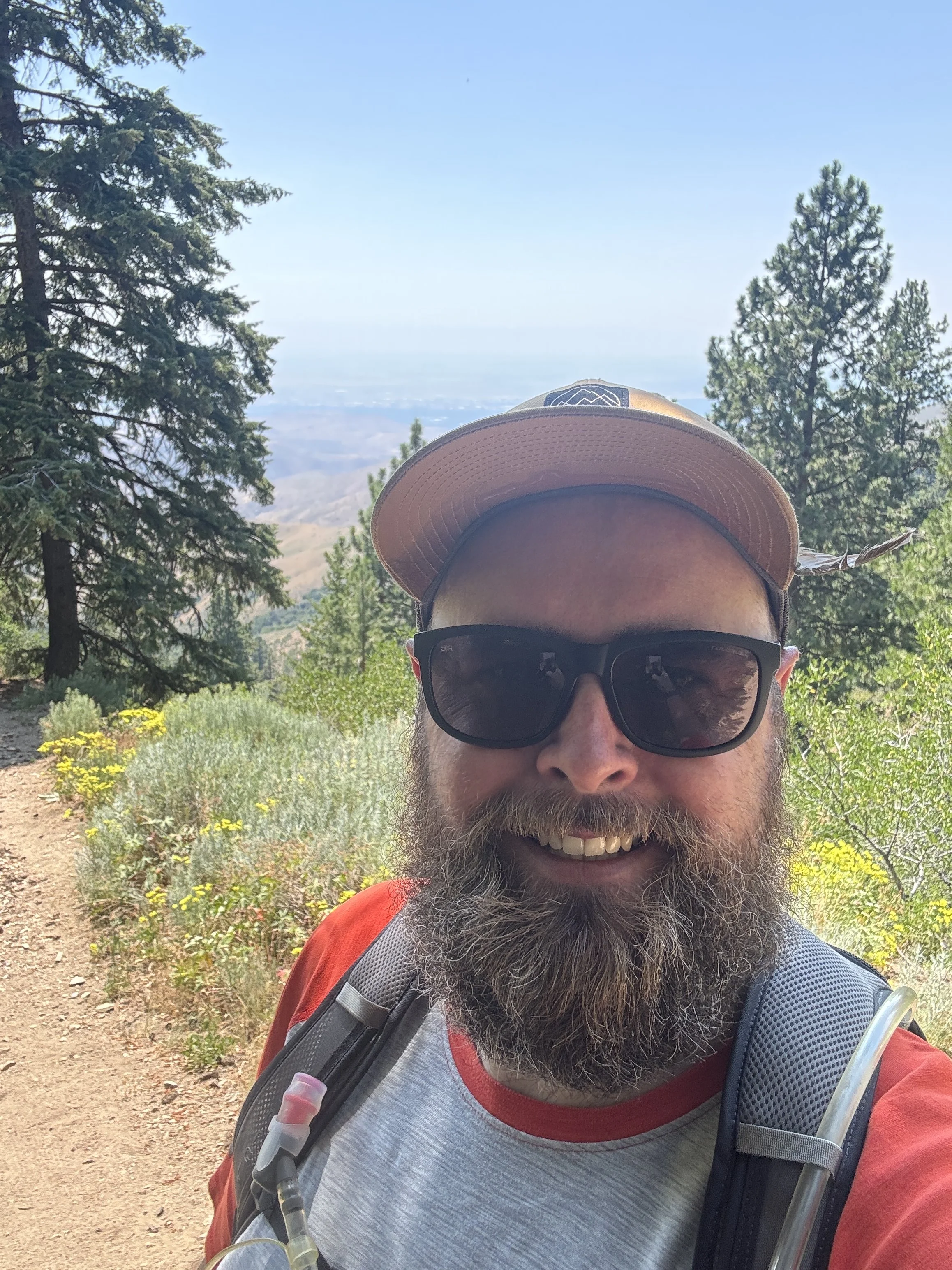 A man with a beard taking a selfie on a hiking trail in a forested area with pine trees, wildflowers, and mountains in the background under a clear blue sky.