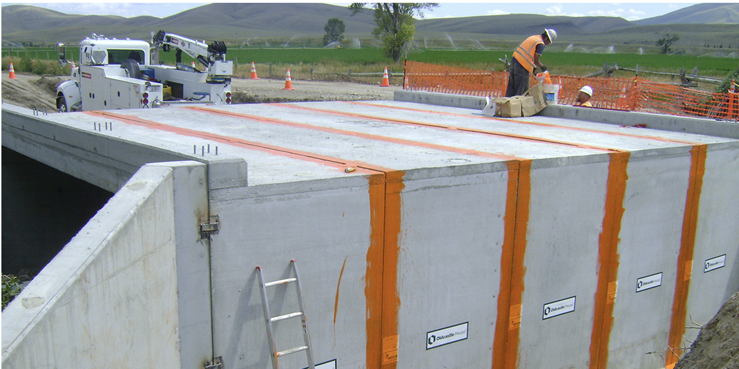 Construction workers working on a bridge deck with orange safety markings, and a utility truck parked nearby, in a rural area with hills and green fields.