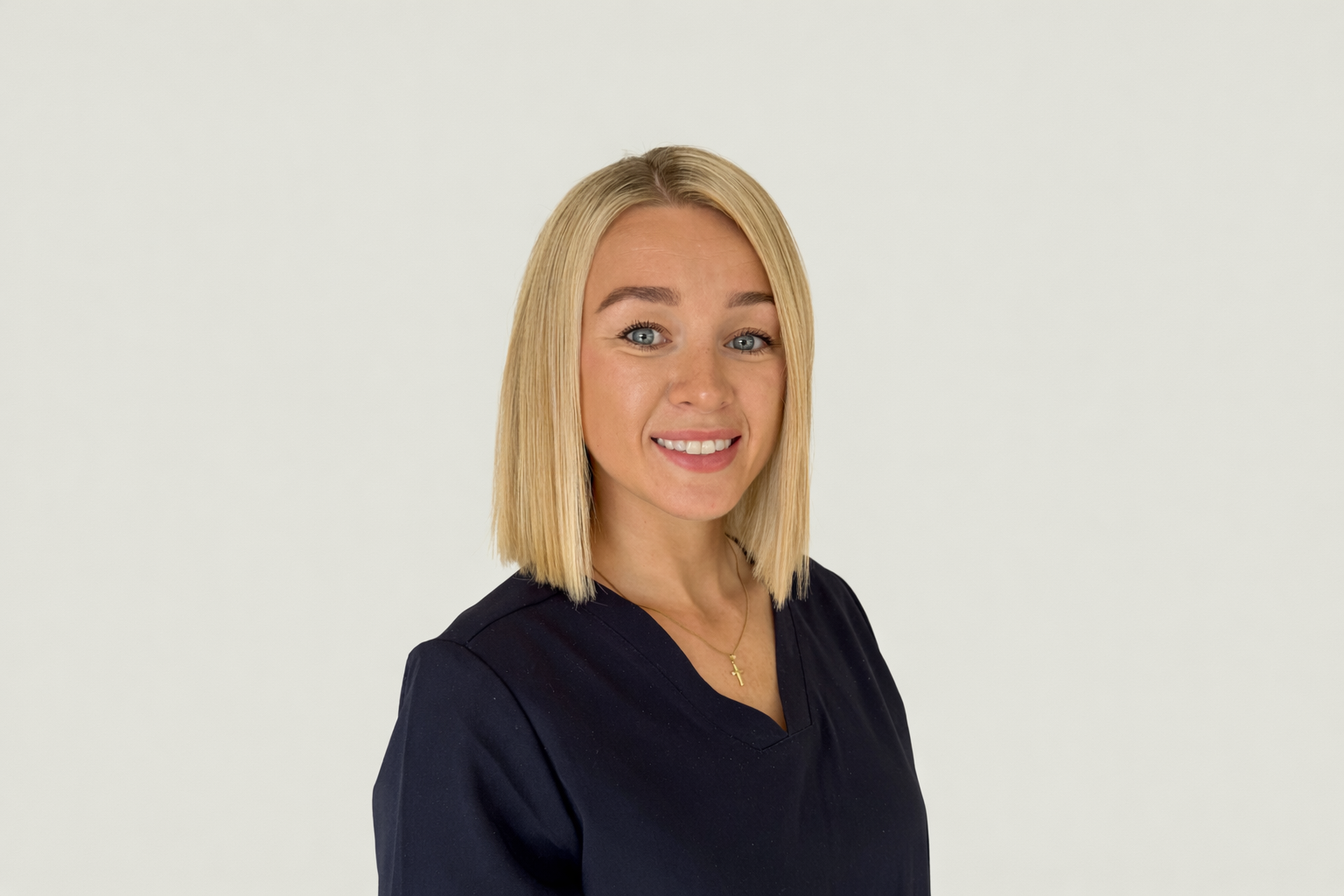 Portrait of a young woman with straight blonde hair, wearing a black top and a gold necklace with a cross pendant, smiling against a plain white background.