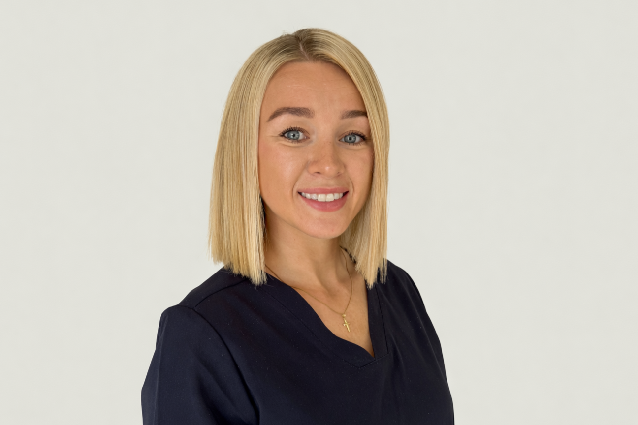 A young woman with blonde hair, blue eyes, and a friendly expression, wearing a black top and a small gold cross necklace, standing against a plain white background.