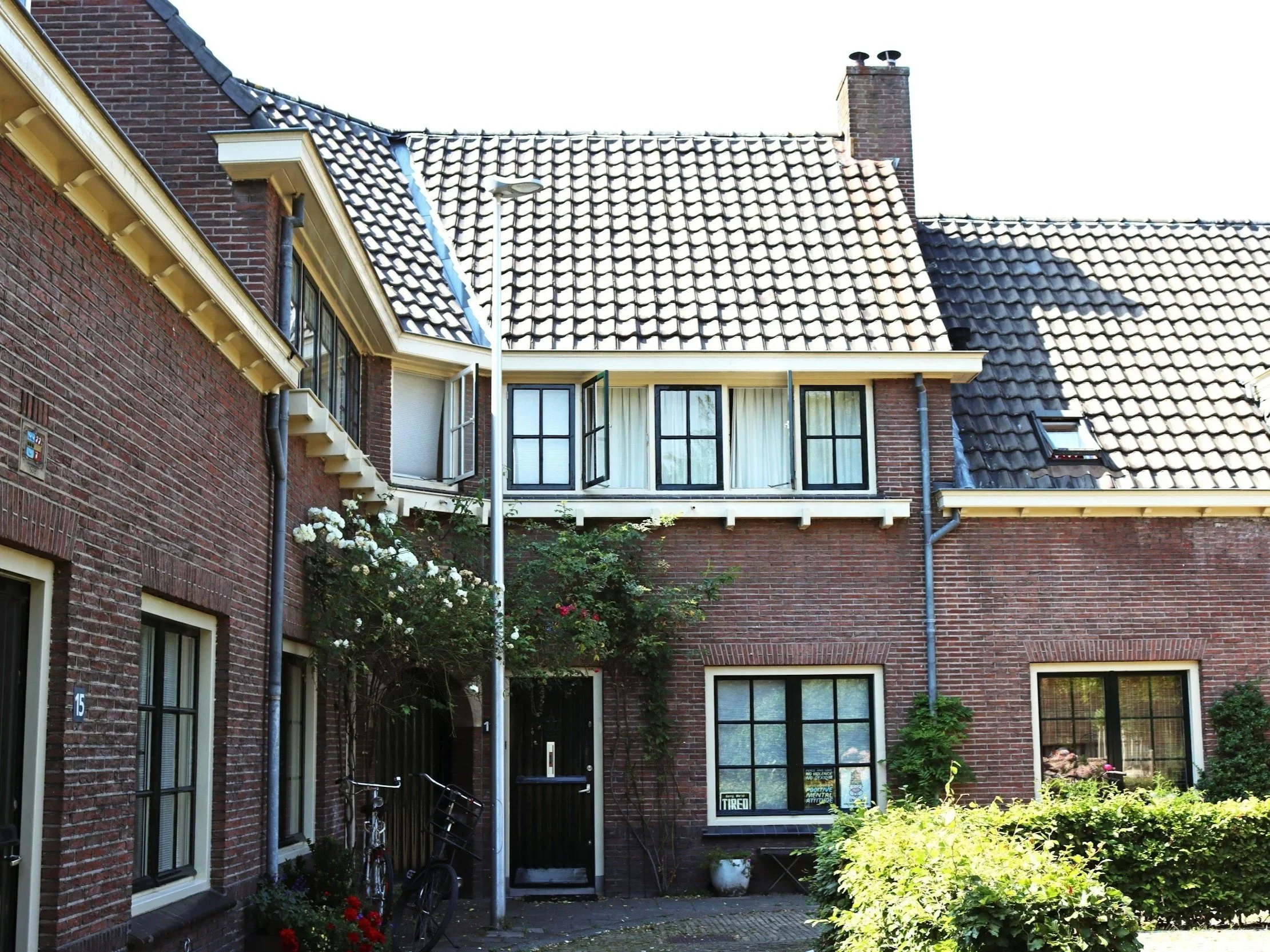 Brick residential building with multiple windows, some with black frames, and a tiled sloped roof with skylights. There is greenery with bushes and flowers in front, and bicycles parked near the entrance.