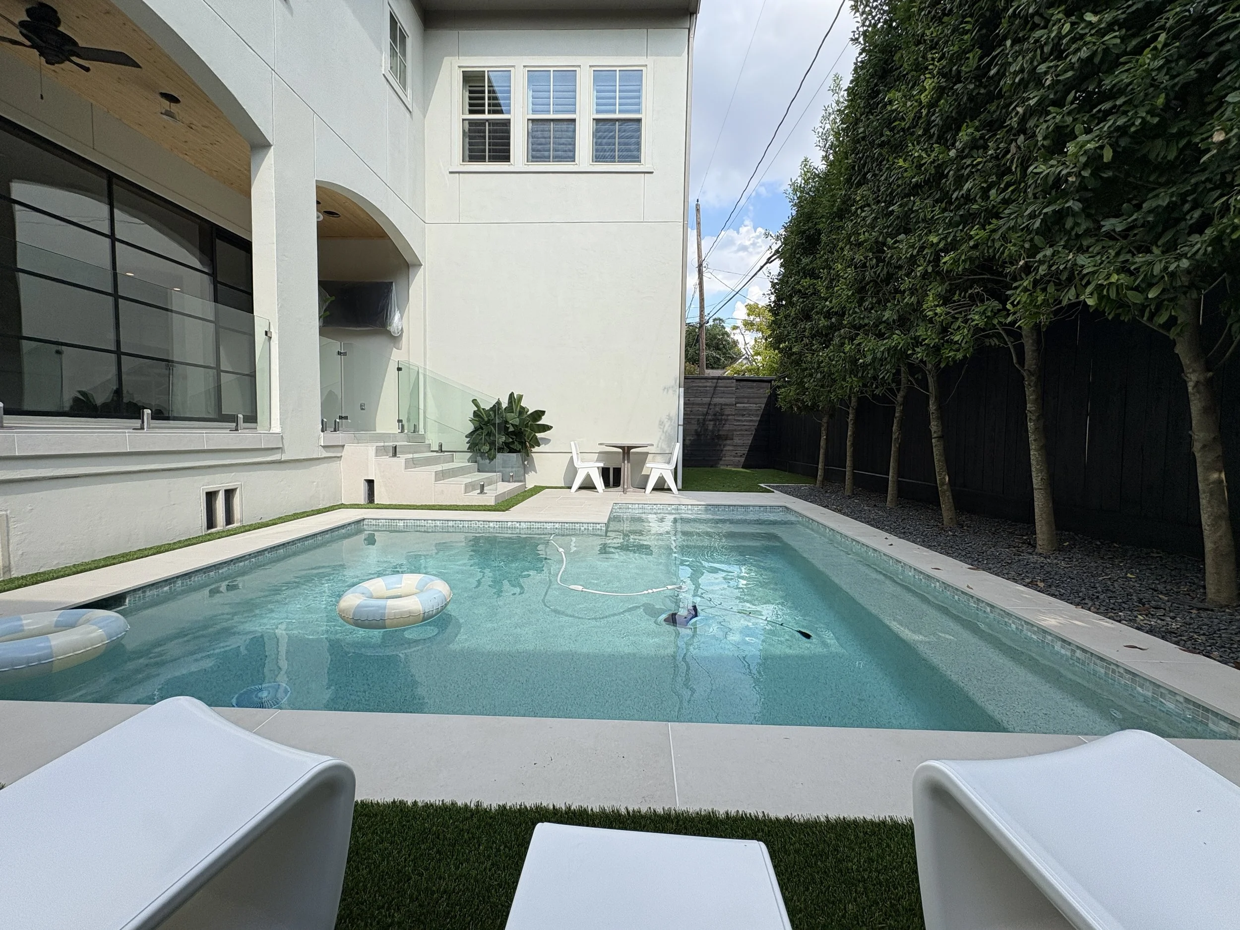 A backyard swimming pool with two inflatable floats and a pool cleaning device in the water. In the background, there's a modern white house with large windows, a small table with two white chairs, some potted plants, and a row of trees along a black