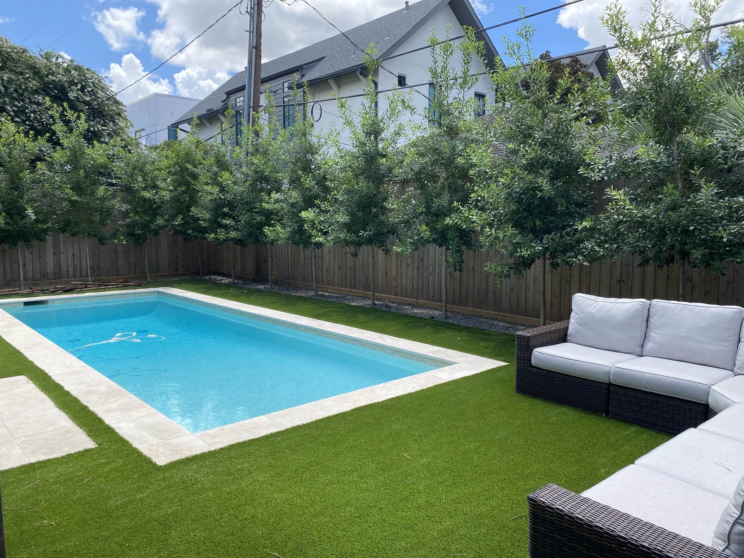 A backyard with a rectangular swimming pool, surrounded by artificial grass and a wooden fence. There are grey outdoor sofas on the right, with trees and a house in the background under a partly cloudy sky.