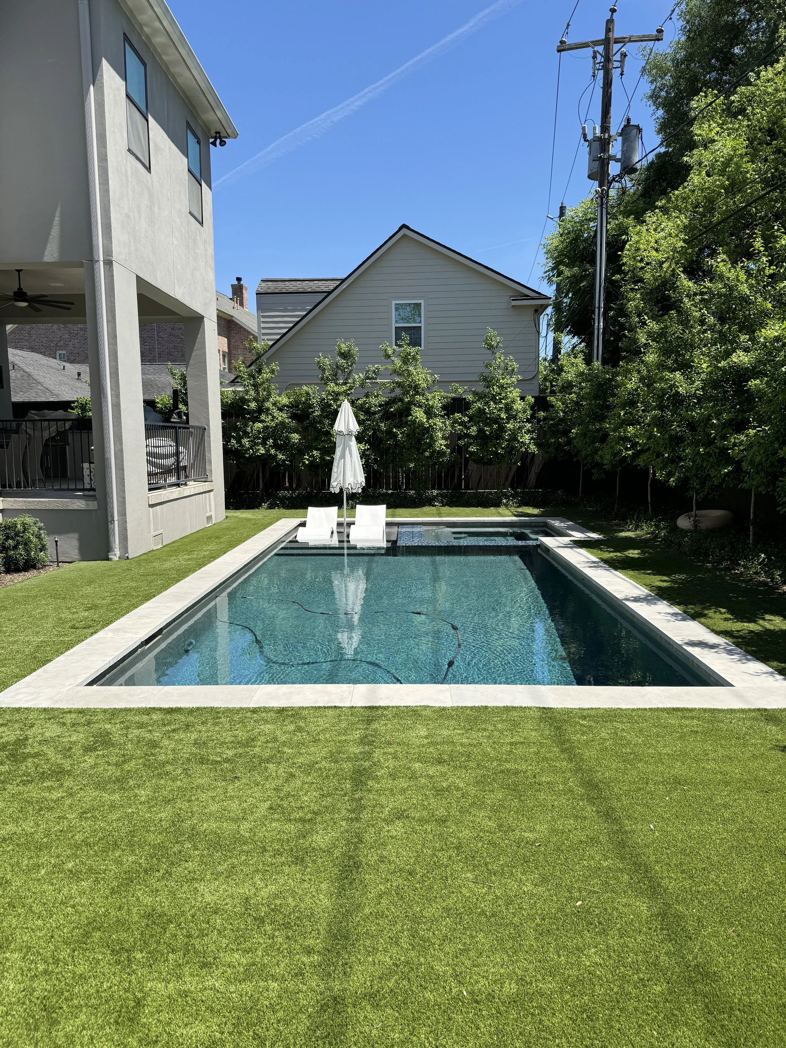 A backyard with a rectangular swimming pool, green grass, two white lounge chairs with an umbrella, houses, trees, and a blue sky.