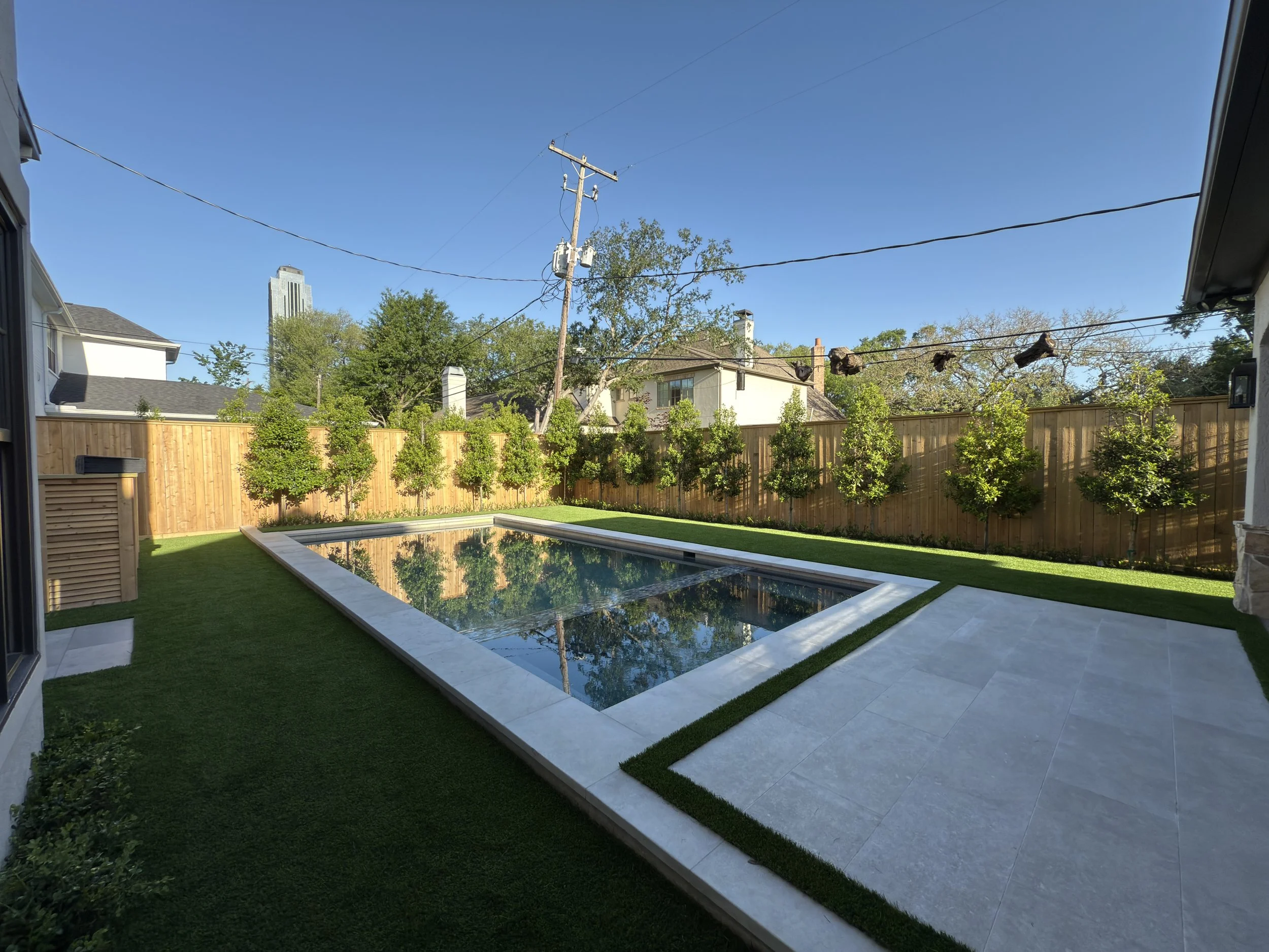 Backyard with a swimming pool, surrounded by a wooden fence, with trees and a house in the background, and a clear blue sky.