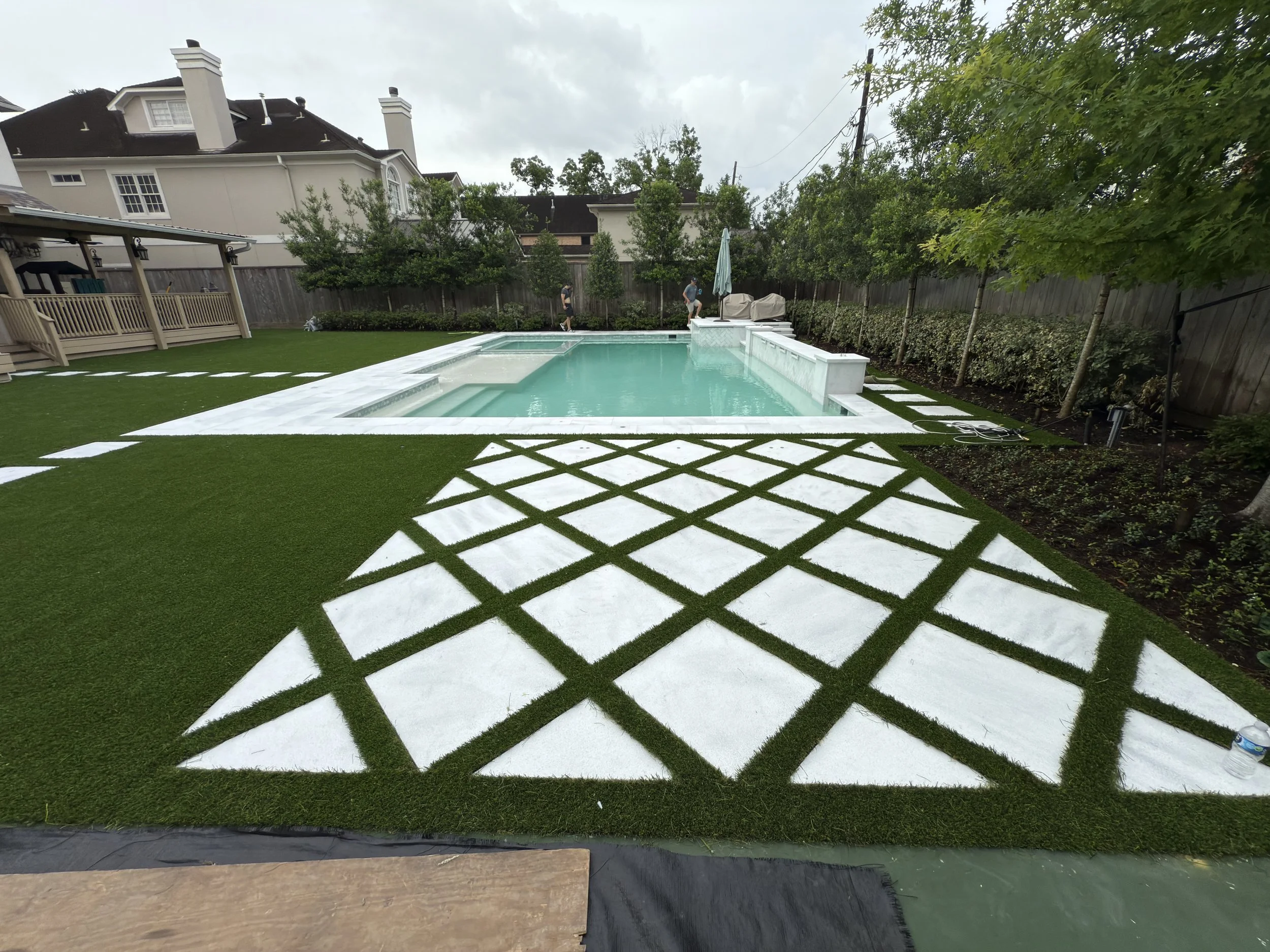 Backyard with a pool, adjacent to a geometric patterned area with white and green grass, surrounded by a wooden fence, trees, and neighboring houses.