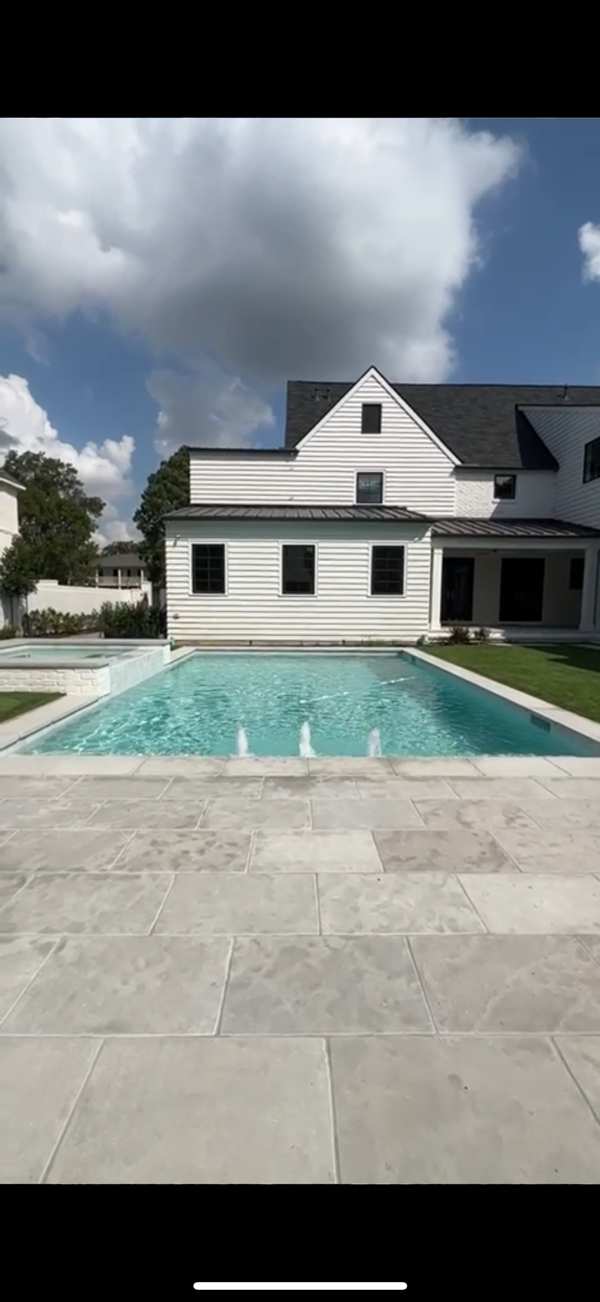 A backyard with a swimming pool, a white house with black shutters, and a grassy area, under a sky with clouds.