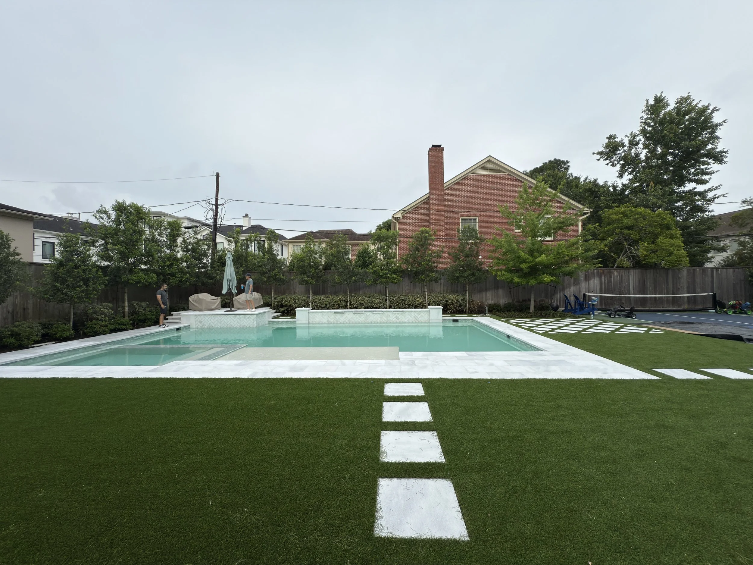 Backyard with a swimming pool, green grass, and a brick house in the background, with tall trees surrounding the yard.