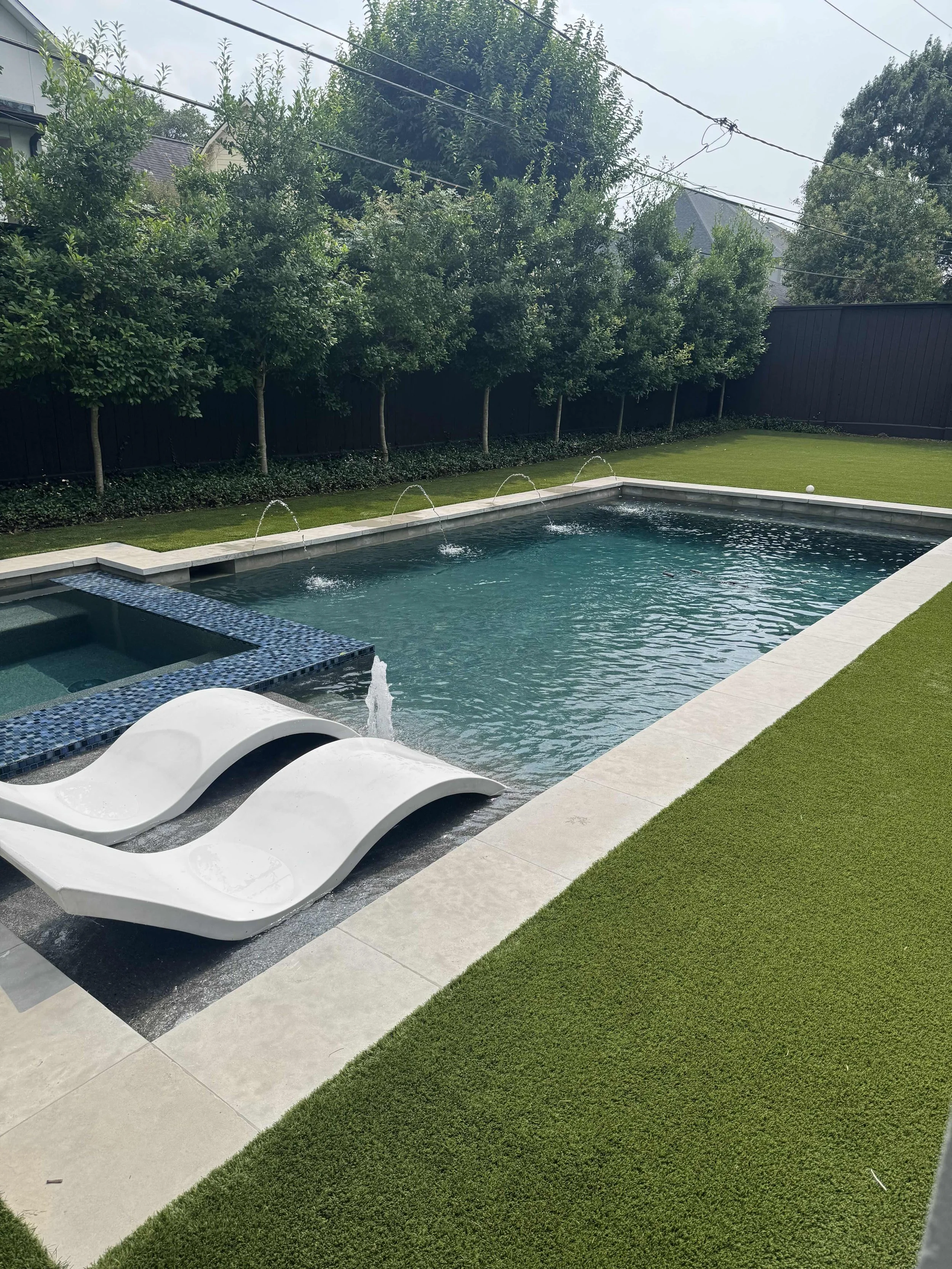 Backyard swimming pool with water fountain jets, a small water slide, and a hot tub surrounded by green lawn and a black fence, with trees and houses in the background.
