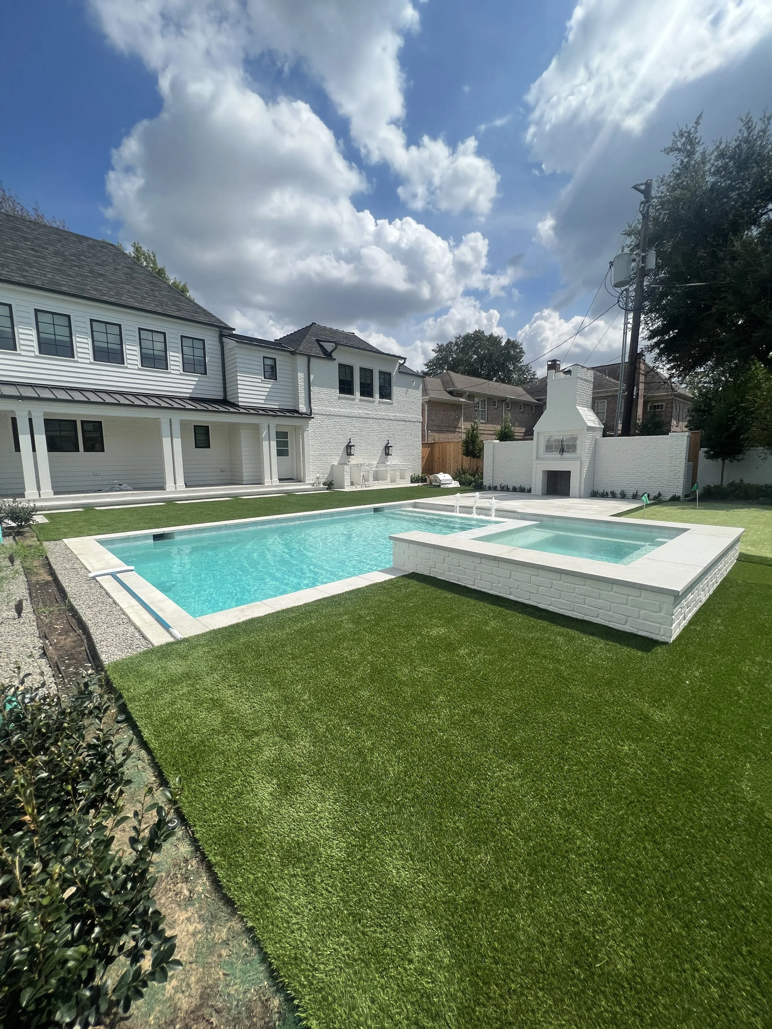 A backyard with a swimming pool and a hot tub, surrounded by green grass and a white brick wall. Modern house and outdoor fireplace are visible in the background, with a partly cloudy sky overhead.
