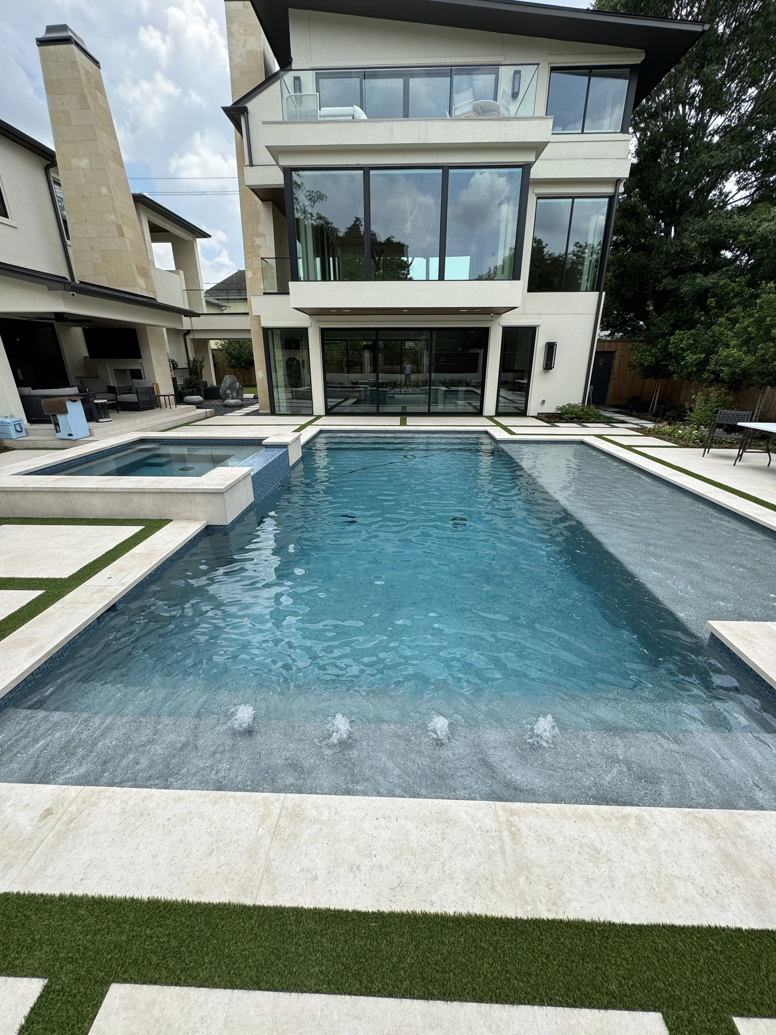 Modern house with large glass windows overlooking a backyard pool with built-in fountains, surrounded by a tiled patio and grass strips, under a partly cloudy sky.