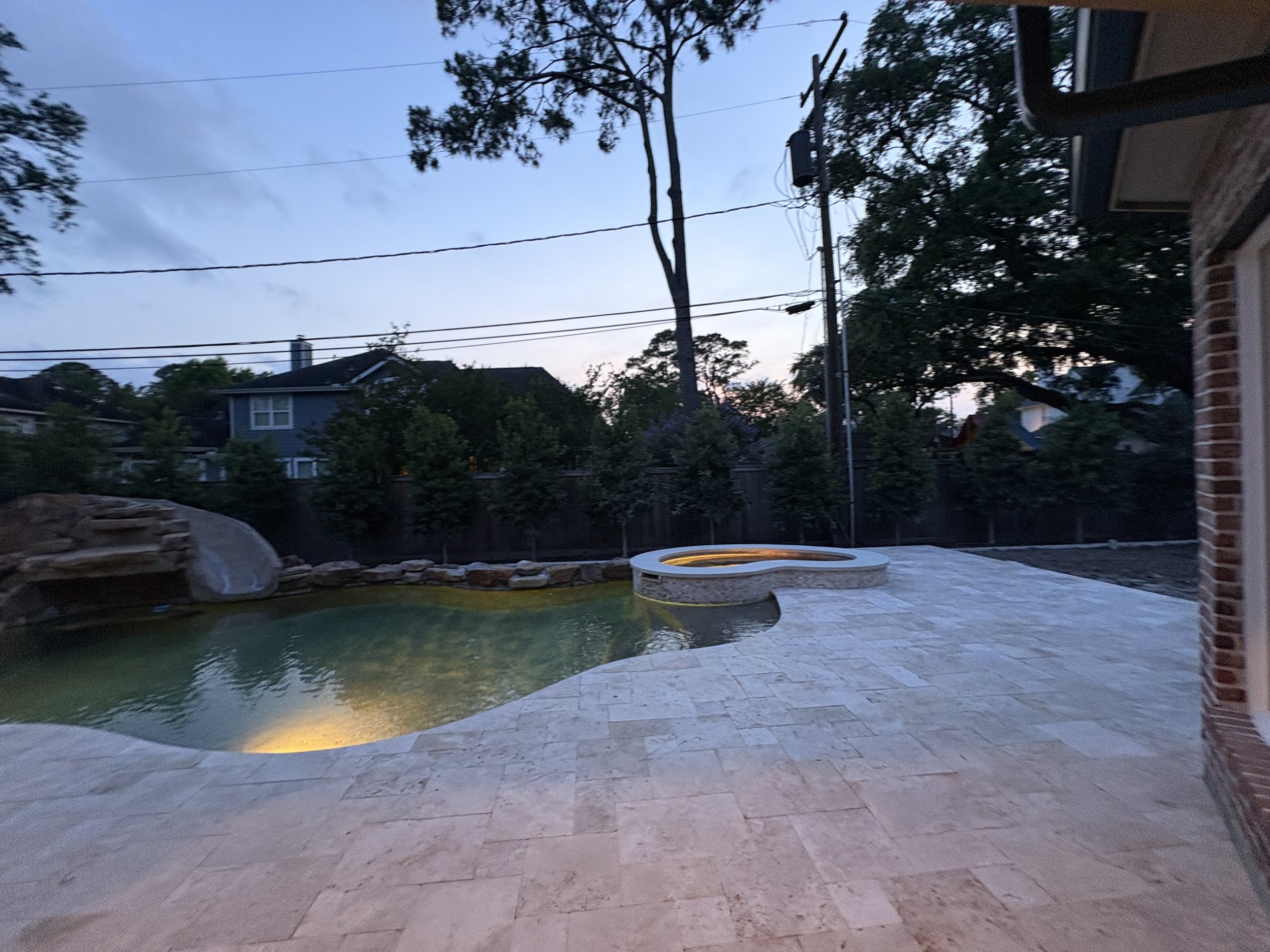 Swimming pool and hot tub in backyard at dusk with trees and houses in the background.