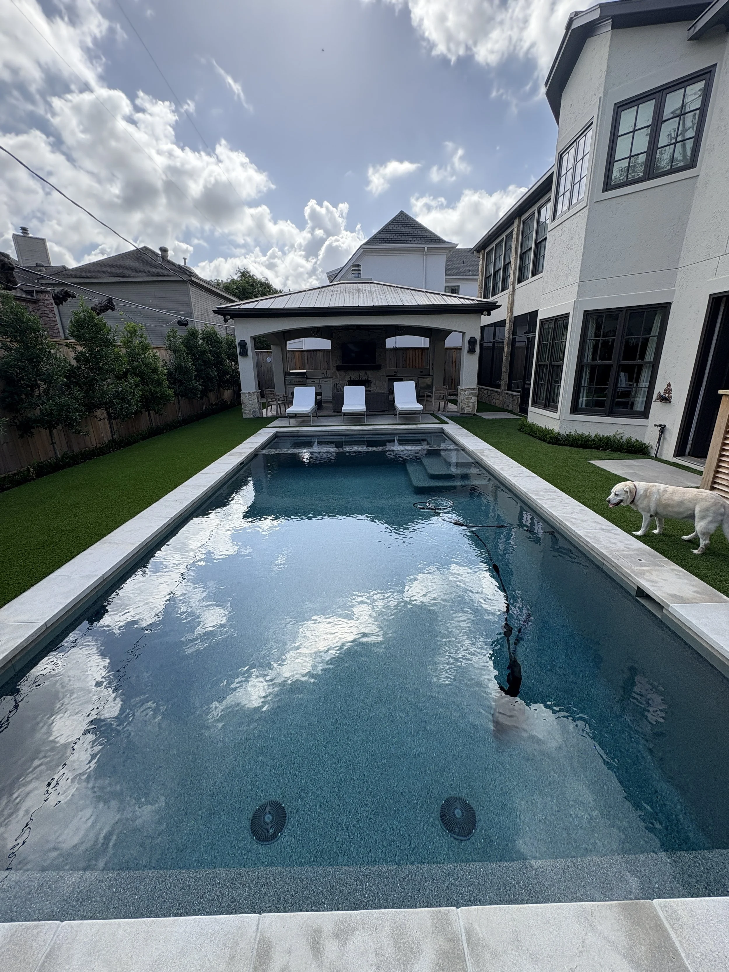 Backyard swimming pool with three white lounge chairs, a covered outdoor kitchen area, and a Labrador retriever dog on the grass next to the pool, with a two-story house and a cloudy sky in the background.