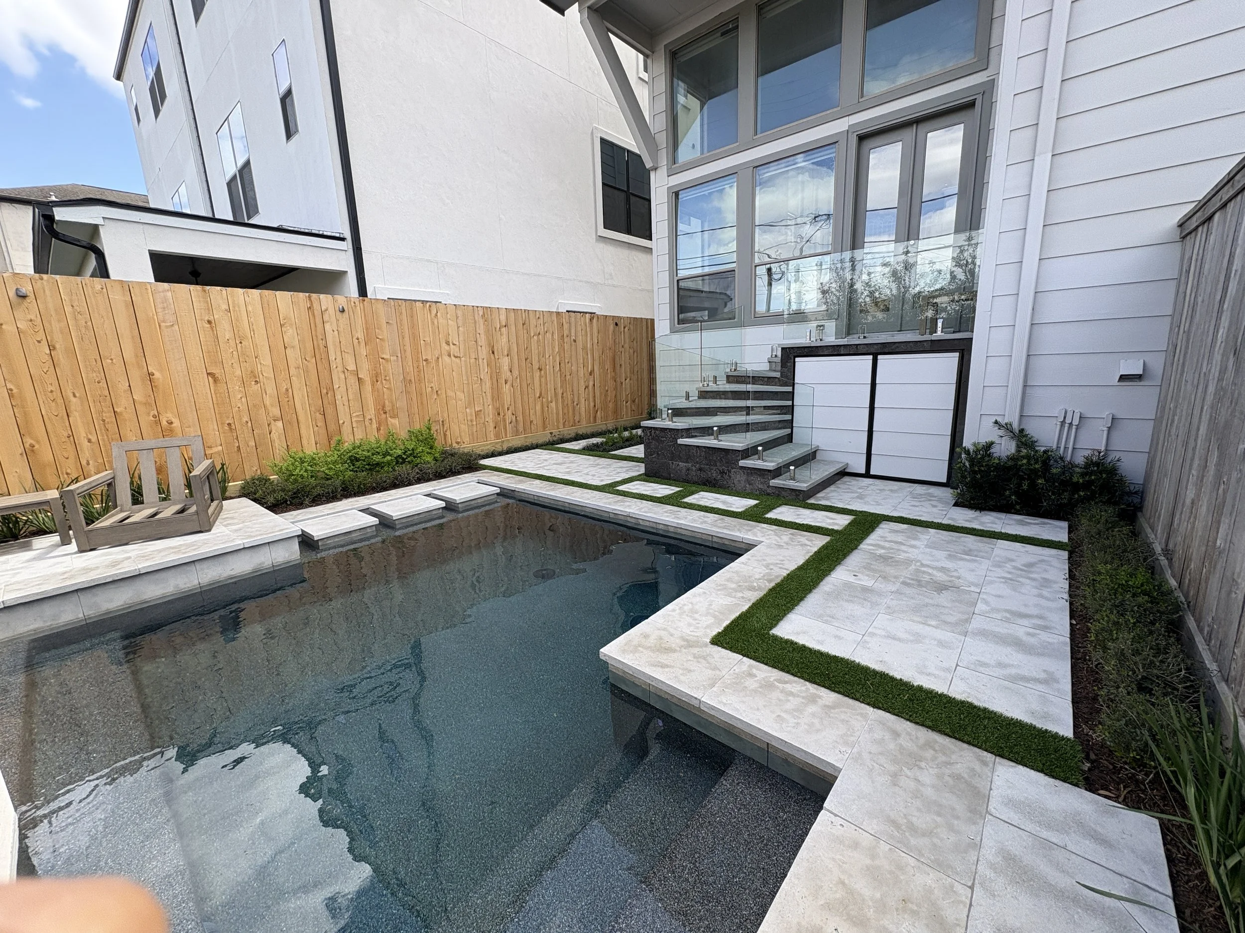 Modern backyard with a small rectangular pool, stone pathway, a white house with large windows, a wooden fence, and small garden plants.