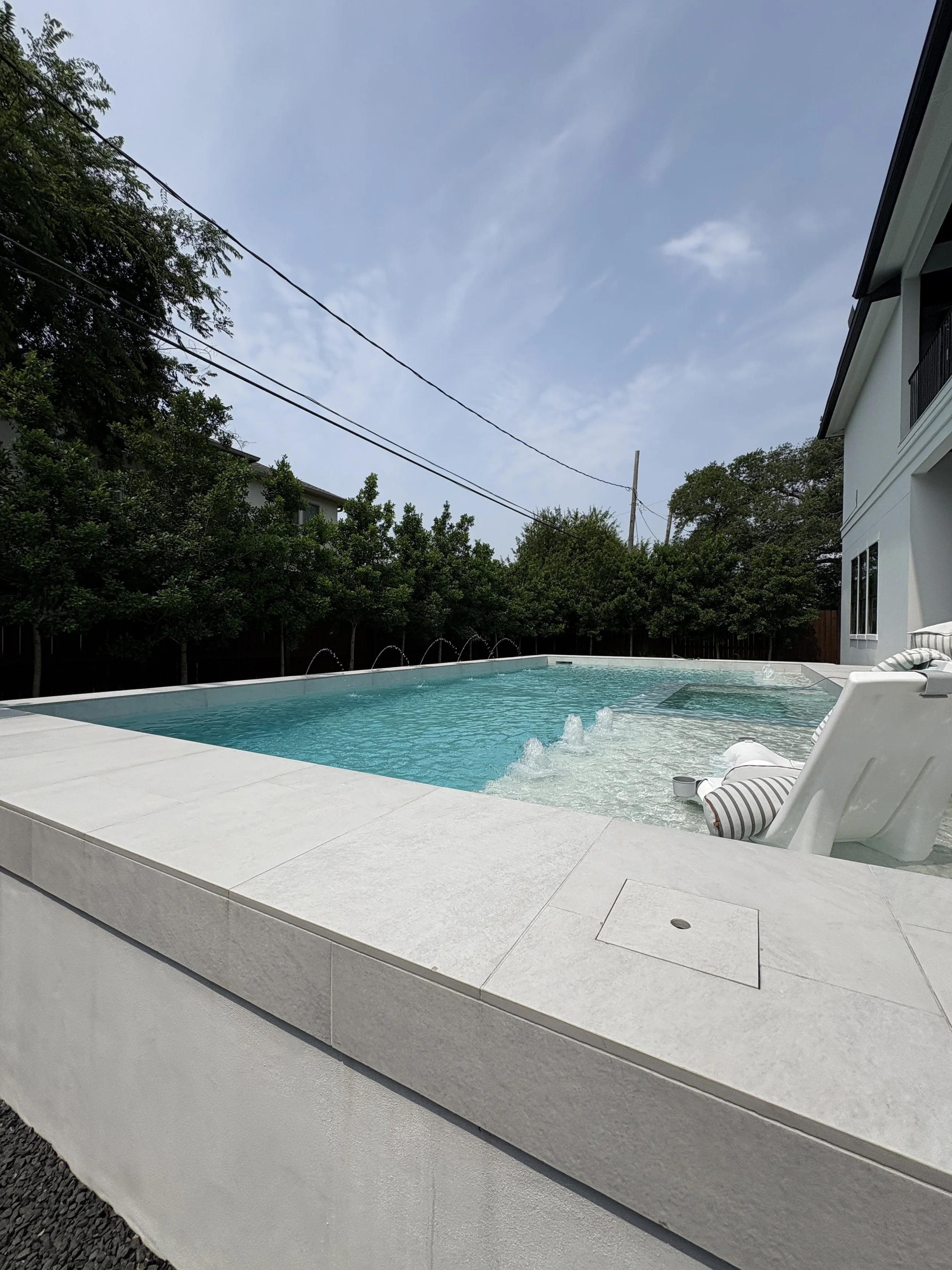 A backyard swimming pool with clear blue water and bubbling fountains, surrounded by a white stone deck, with a lounge chair and striped pillow on the right side, and greenery and trees in the background under a cloudy sky.