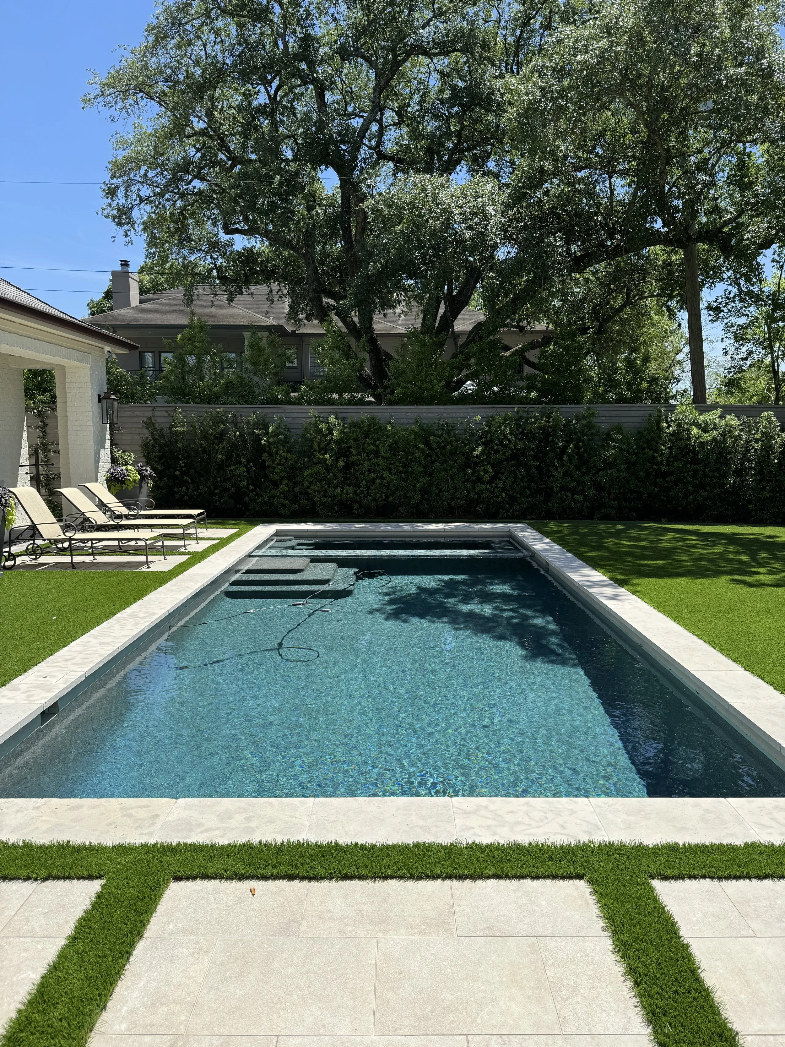 A rectangular swimming pool in a backyard with a hot tub at the far end, surrounded by green grass, a white tiled patio, and four lounge chairs. Tall trees and a house with a dark roof are in the background under a clear blue sky.