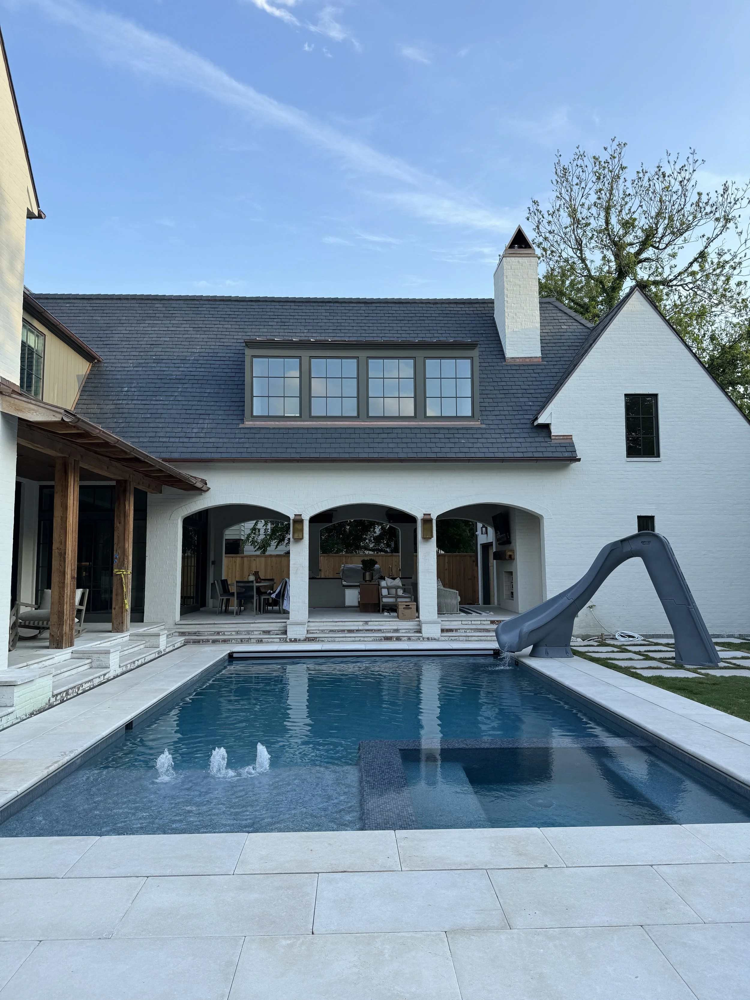 Backyard with a swimming pool, water slide, covered patio with outdoor furniture, and a two-story white house with large windows and a chimney.
