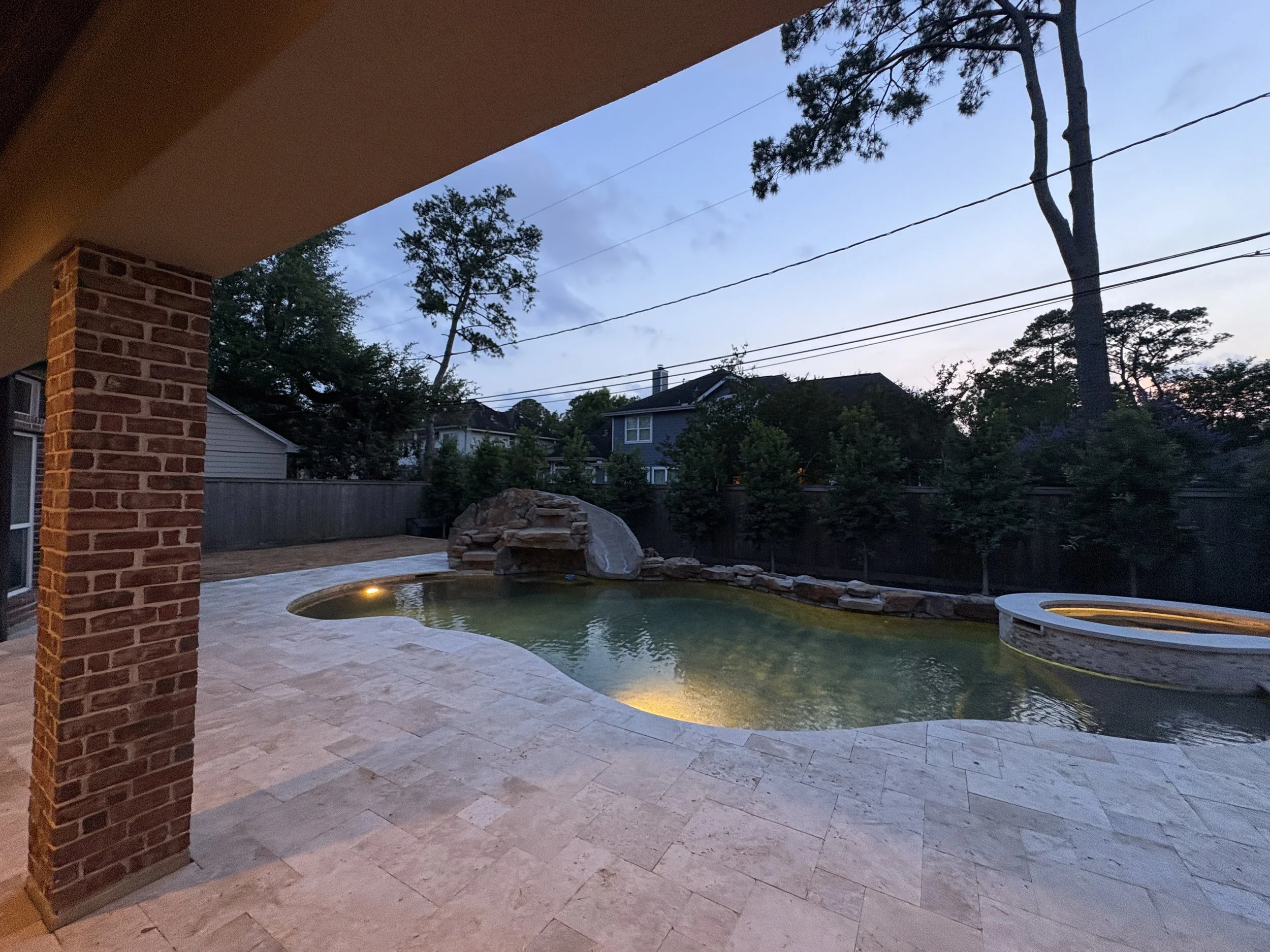 Backyard with a swimming pool and hot tub at dusk, brick patio, trees, and neighboring houses in the background.