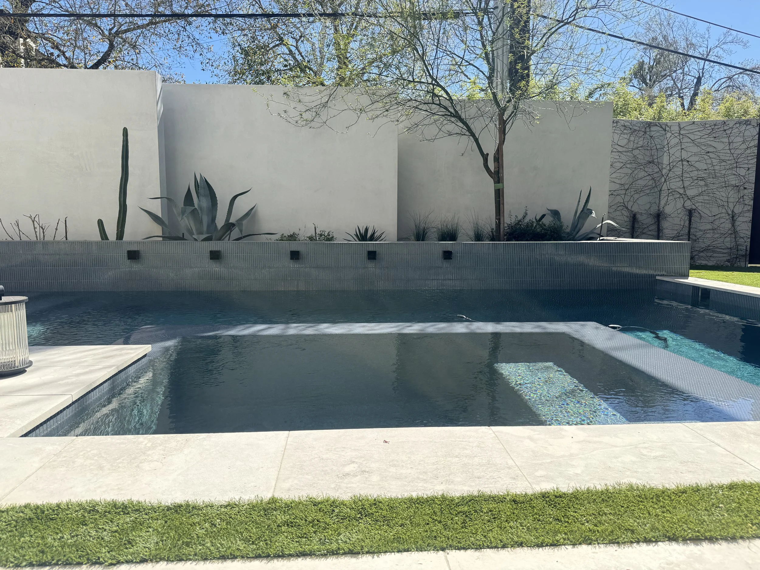 A modern backyard swimming pool with a spa, surrounded by a concrete deck and a white privacy wall with desert plants and trees.