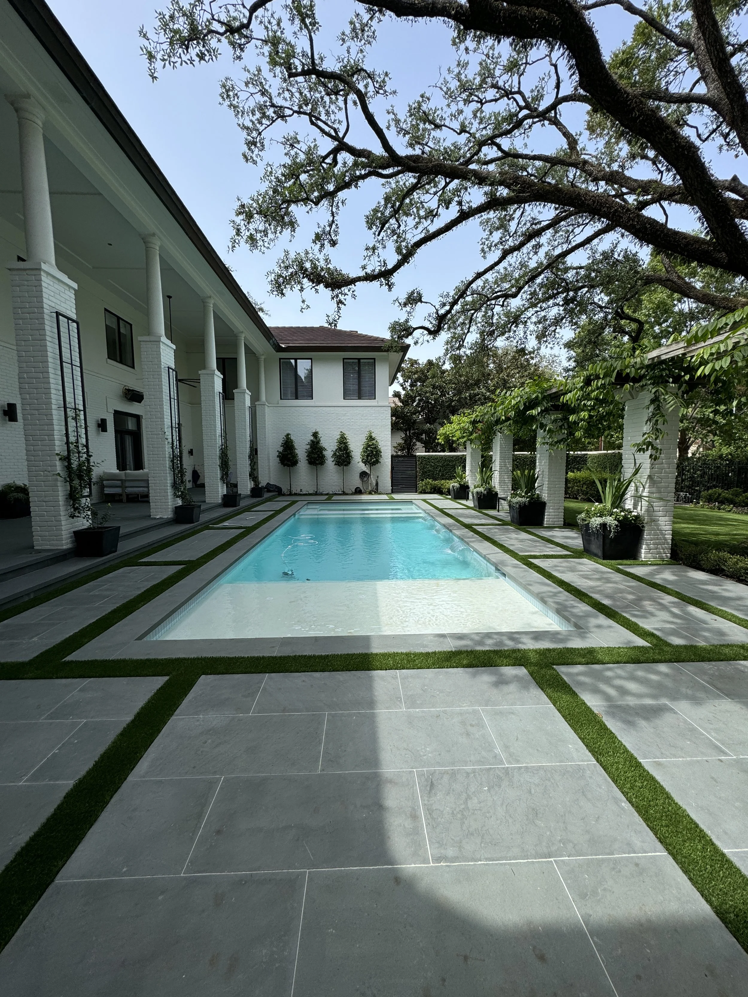 Modern backyard with a rectangular swimming pool, grey tiled patio with green grass in between, surrounded by white brick columns and house, lush trees, and potted plants.
