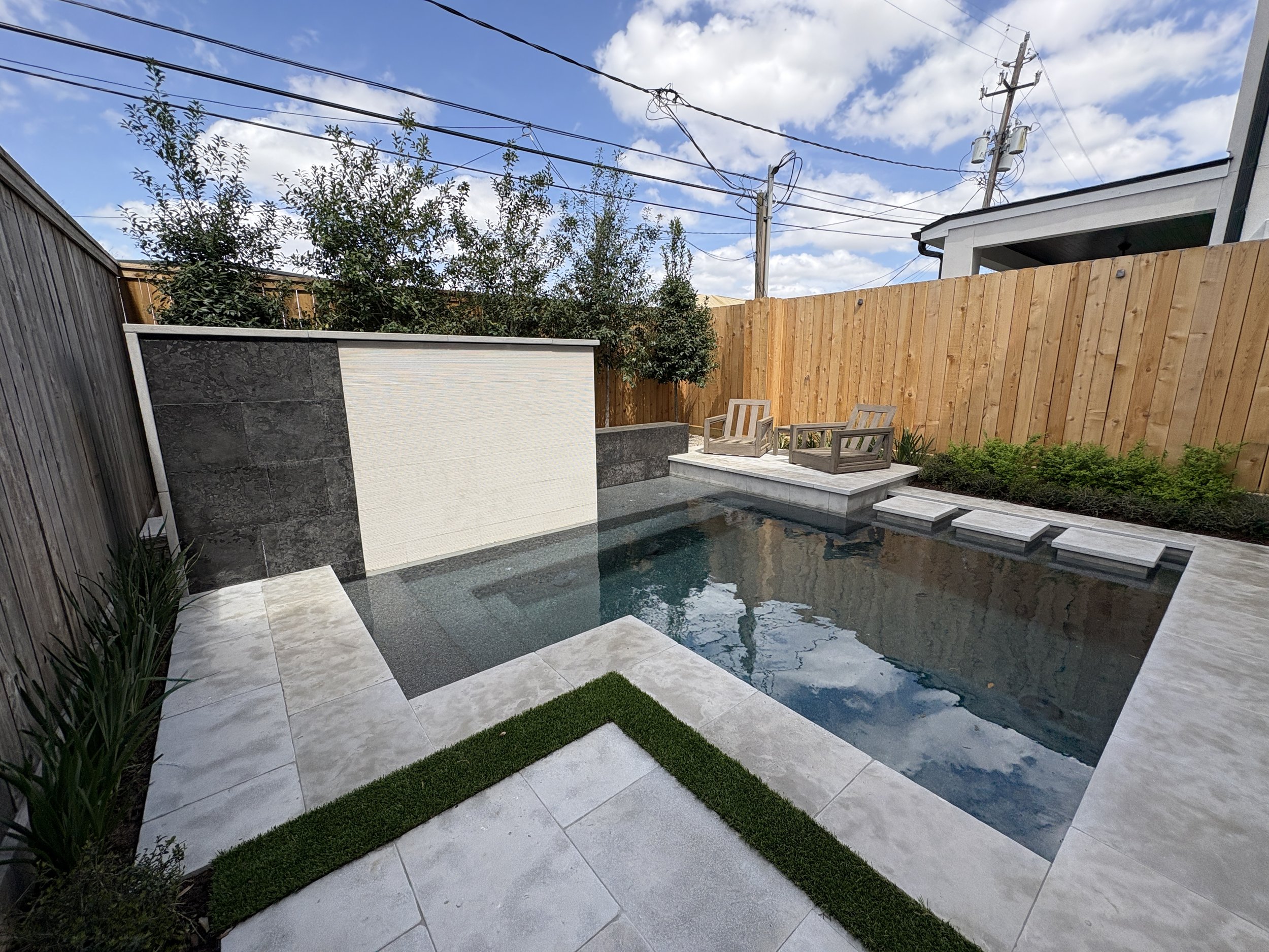 A backyard with a small, empty swimming pool, stone steps leading into it, and a wooden fence. There are two chairs on a small platform at the far end of the pool, plants along the fence, and a cloudy sky overhead.