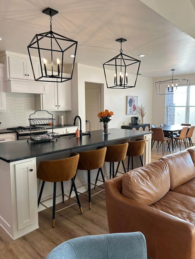 Open-concept kitchen and dining area with black pendant lights, white cabinetry, dark countertop, bar stools, a brown leather couch, and a dining table with six chairs.