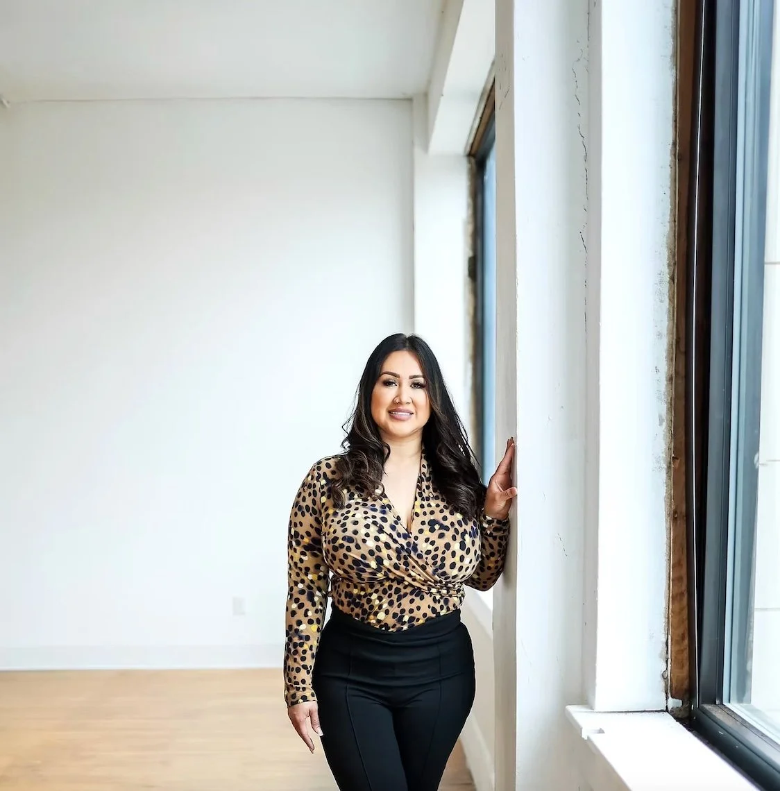 A woman stands near a large window in a spacious, minimally decorated room with white walls and wooden floors, smiling at the camera.