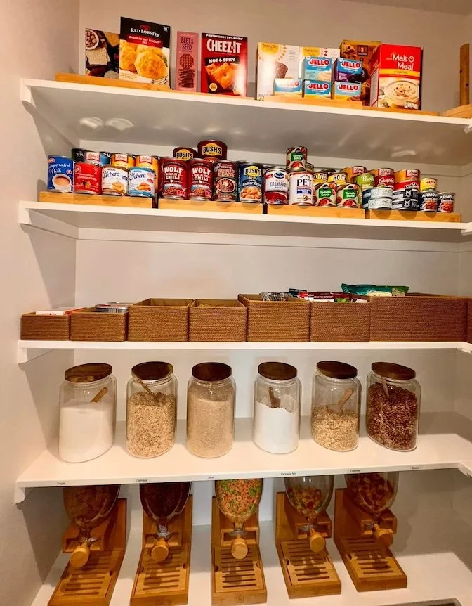 Kitchen pantry shelf with canned foods, boxed meals, jars of grains and spices, and cereal dispensers.