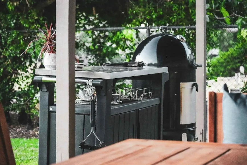 Outdoor patio with a black charcoal grill, a kitchen counter, and a potted plant, surrounded by greenery and a chain-link fence.
