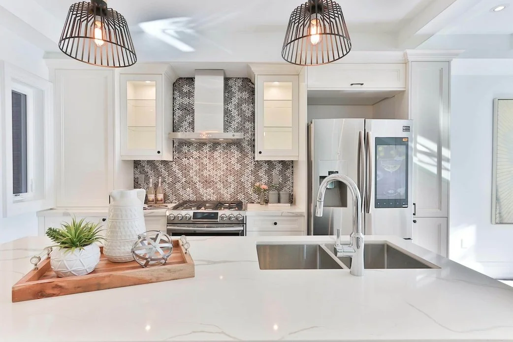 Modern kitchen with white cabinetry, black patterned tile backsplash, stainless steel refrigerator, oven, and range hood, with pendant lights hanging above a white countertop island that has a sink.