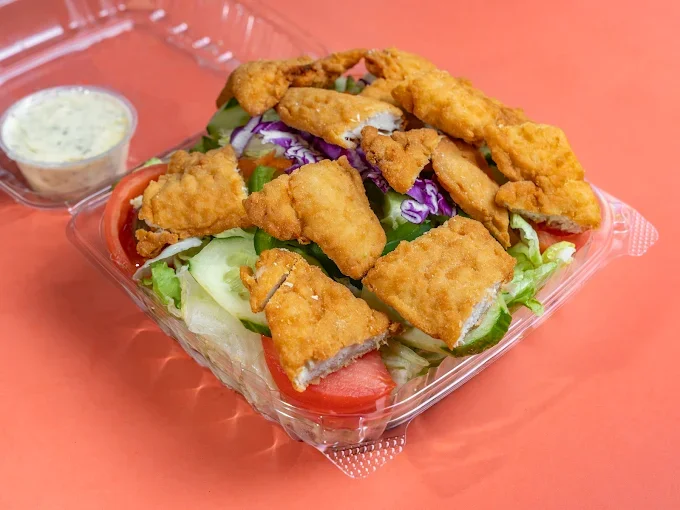 Salad with fried fish fillets, lettuce, tomato, purple cabbage, with a side of dipping sauce on a red table.