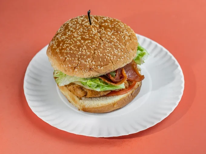A chicken sandwich with lettuce, tomato, bacon, and grilled chicken fillet on a sesame seed bun, served on a white paper plate.