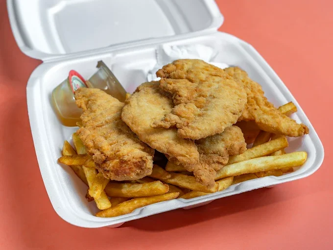 Fried chicken tenders with French fries, a small container of honey mustard sauce, and a packet of ketchup in a white takeout box.