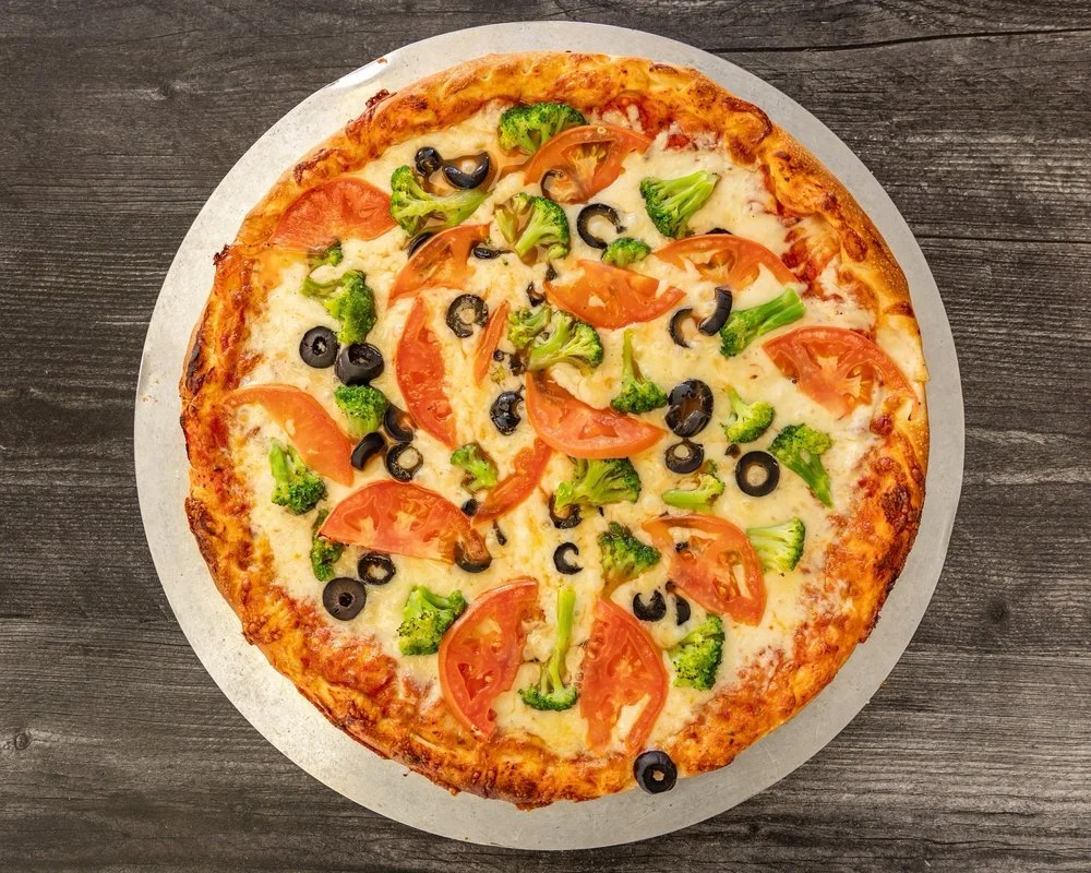 Vegetable pizza topped with tomato slices, broccoli, black olives, and melted cheese on a wooden surface.