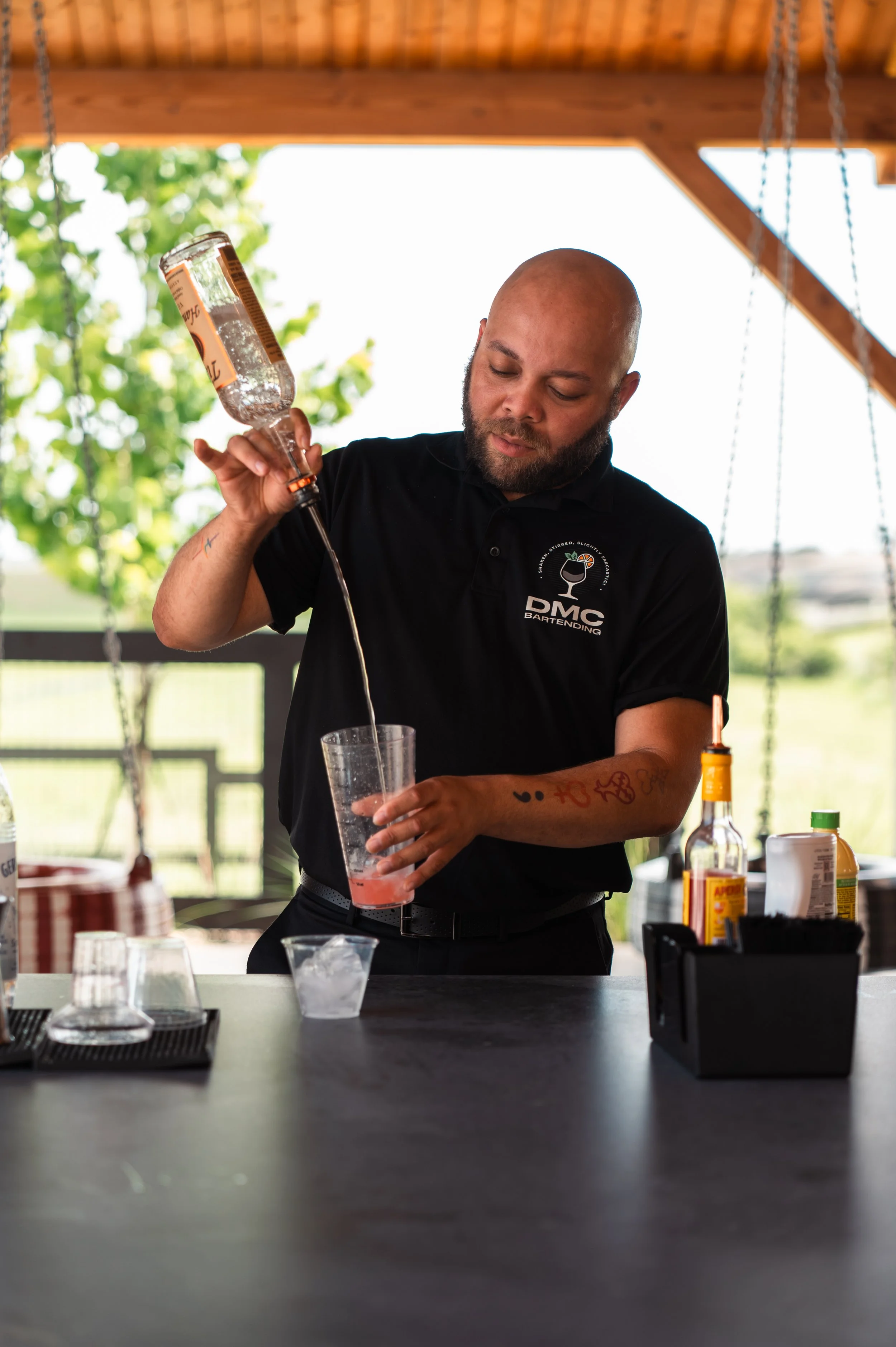 Bartender pouring a pink cocktail into a glass at an outdoor bar with a wooden roof.