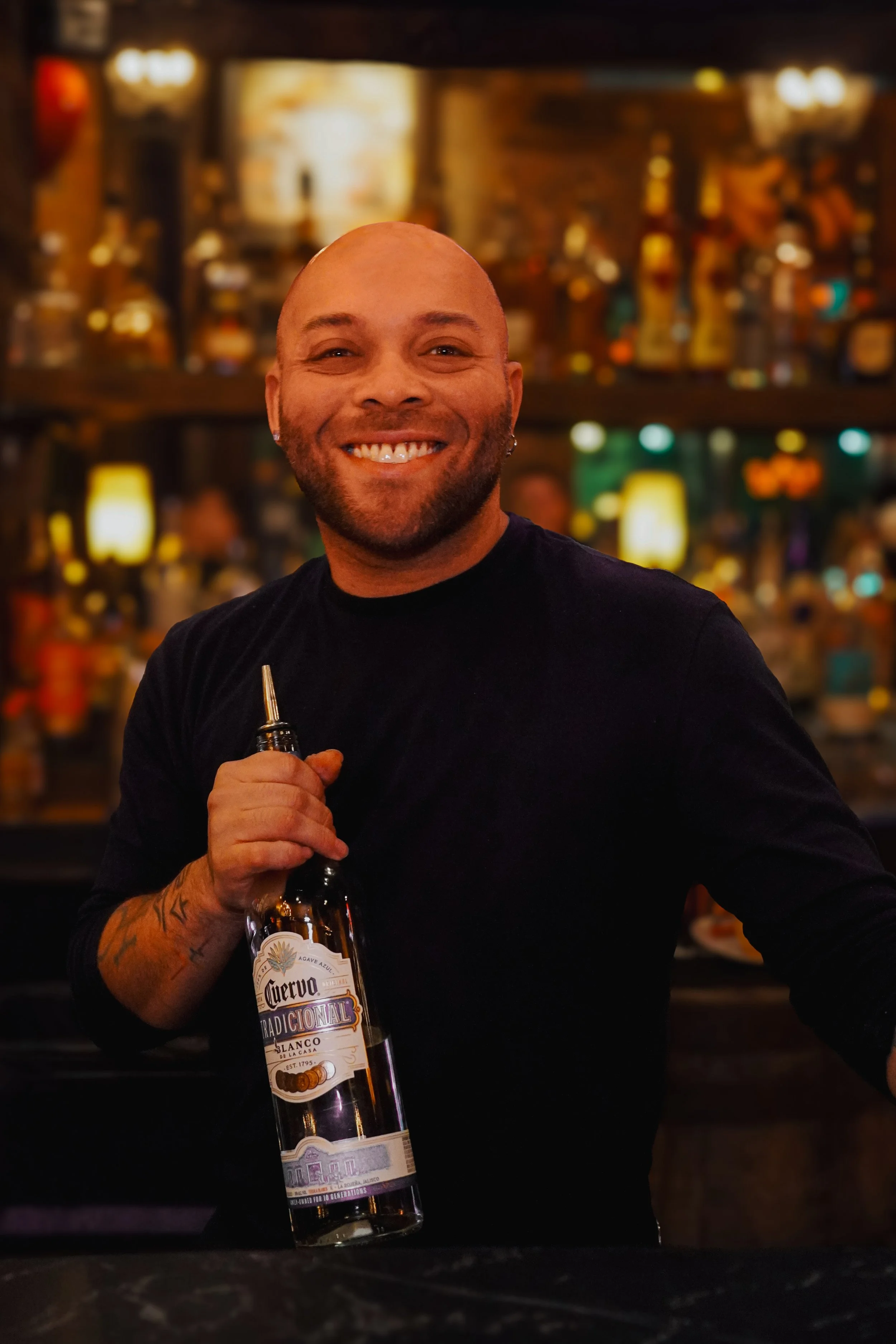 Smiling bartender with tattoos on arm holding a bottle of tequila in a lively bar with warm lighting.