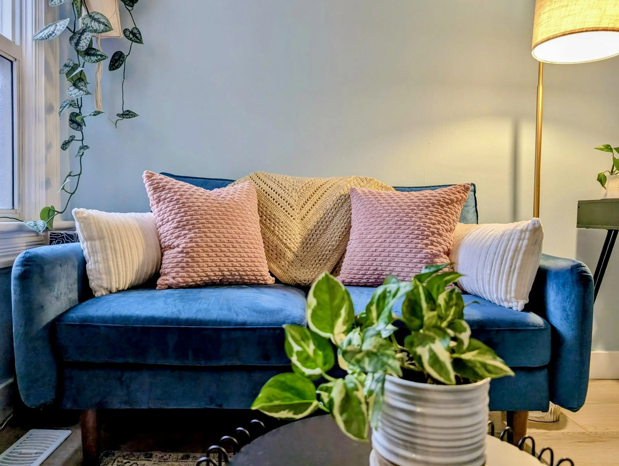 Accord Psychology Office. Therapy office with a blue velvet sofa, pink and white textured pillows, a yellow knitted blanket, a tall yellow floor lamp, and decorative plants on a window ledge and in a white pot.