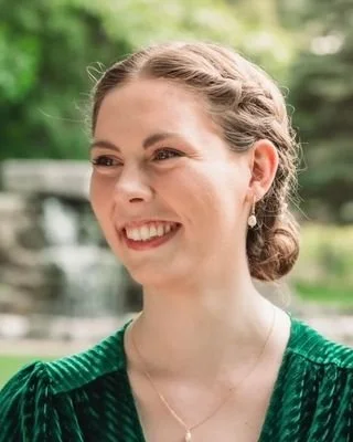 Hannah Majury, Registered Psychotherapist (Qualifying) . A woman smiling outdoors with a green background, wearing a dark green textured top and pearl earrings.