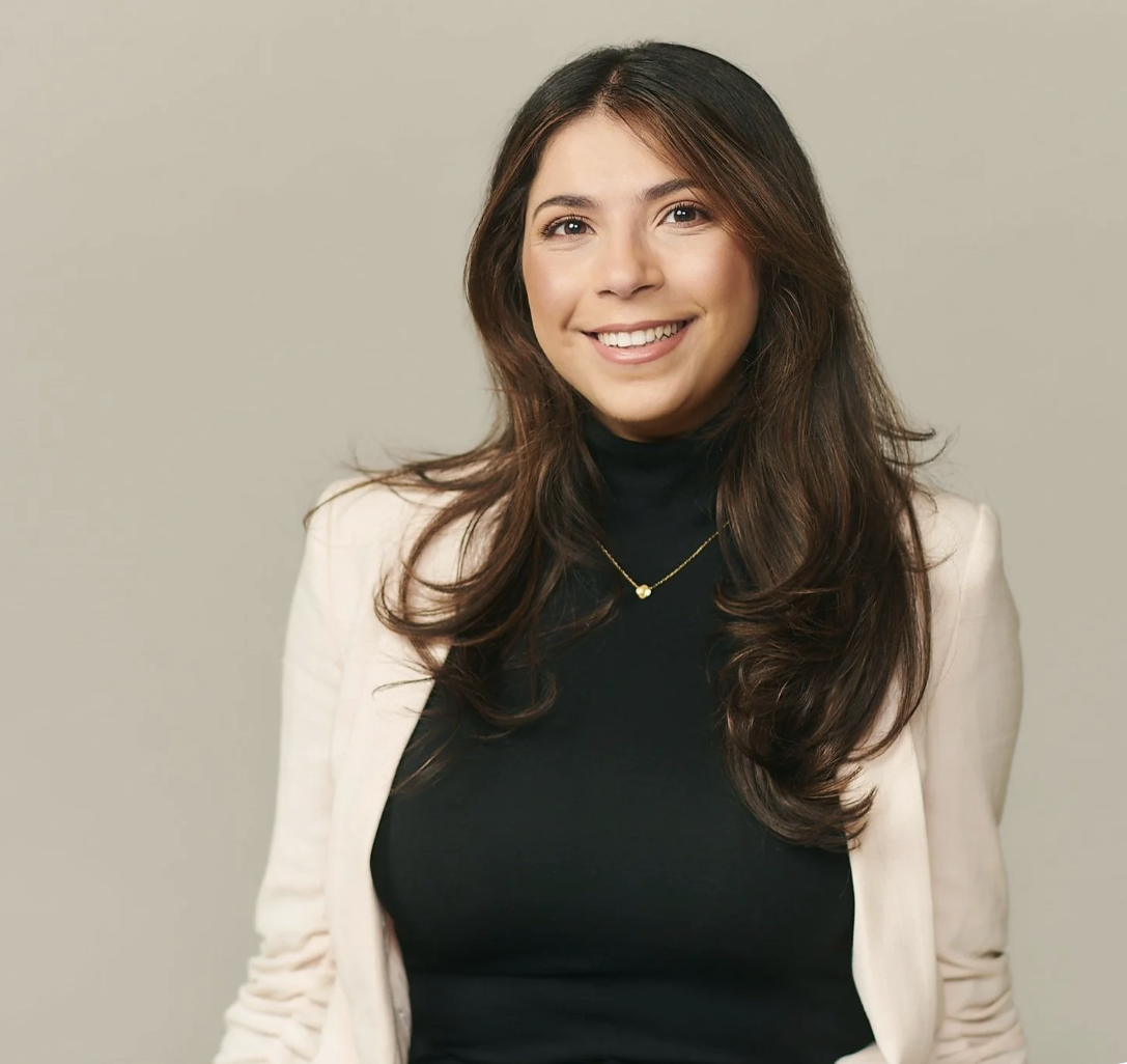 Marwa Ibrahim, Accord Psychology Clinical Assistant. A woman with long brown hair, wearing a cream blazer, black turtleneck, and gold necklace, smiling against a plain background.