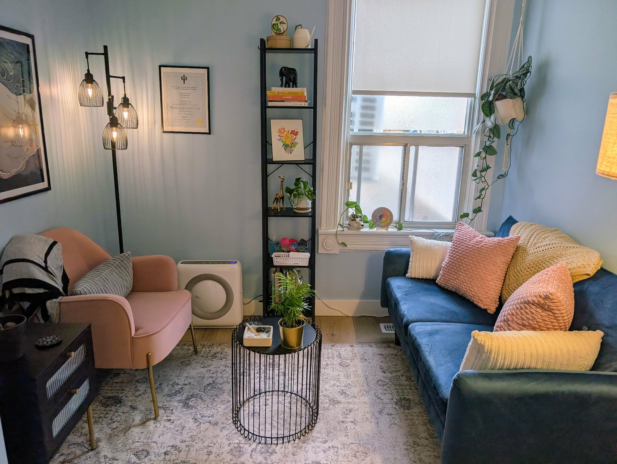 A cozy office with a pink armchair, a black bookshelf, a blue sofa with pink and yellow pillows, a small round black wire coffee table with a plant, a patterned area rug, and a window with a hanging plant on the right.