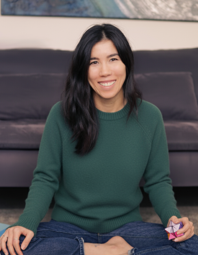 Dr. Melodie Lemay-Gaulin, Clinical Psychologist. A woman with long black hair sitting on the floor in front of a black couch, smiling at the camera, wearing a green sweater and jeans, holding a colorful geometric cube puzzle in her right hand.